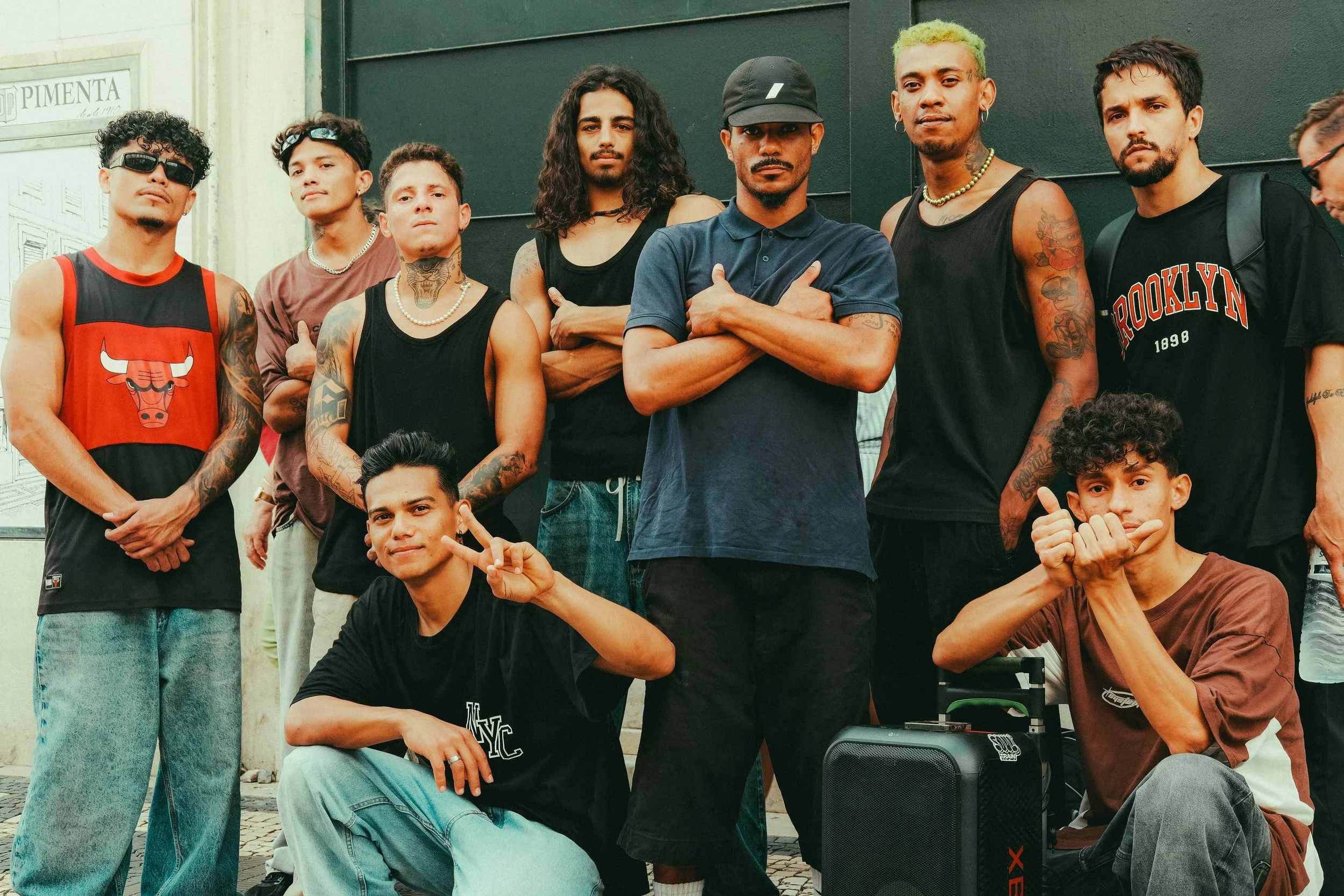 Gruppe junger Menschen posiert draußen vor einer dunklen Wand, einige sitzen, andere stehen, mit verschiedenen Styles und Ausdrücken.