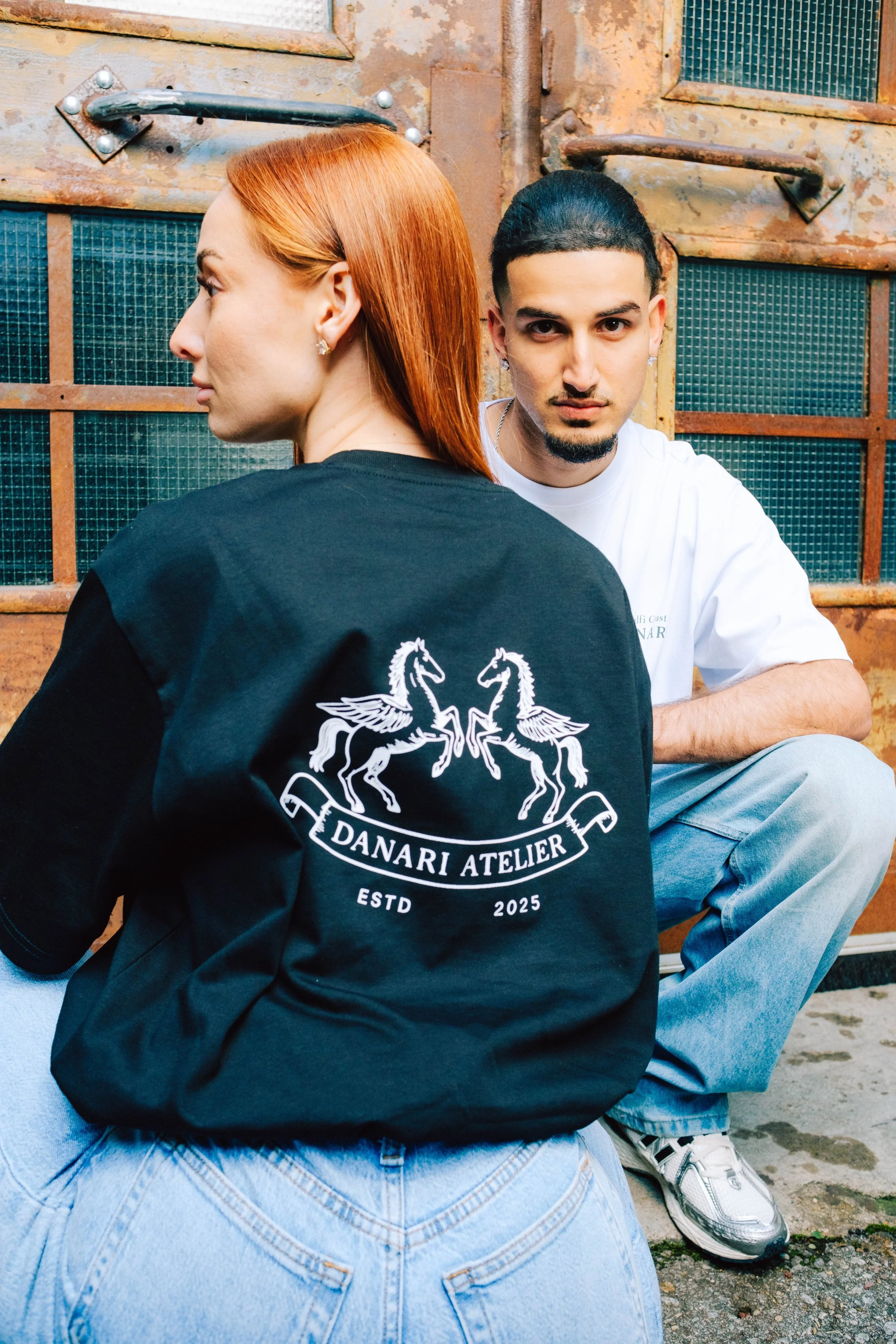 Zwei junge Menschen, ein Mann und eine Frau, sitzen vor einem rusty, alten Tor. Die Frau trägt eine schwarze Jacke mit einem weißen Wappen mit zwei geflügelten Hengsten und dem Text "DANARI ATELIER", während der Mann einen weißen T-Shirt trägt.