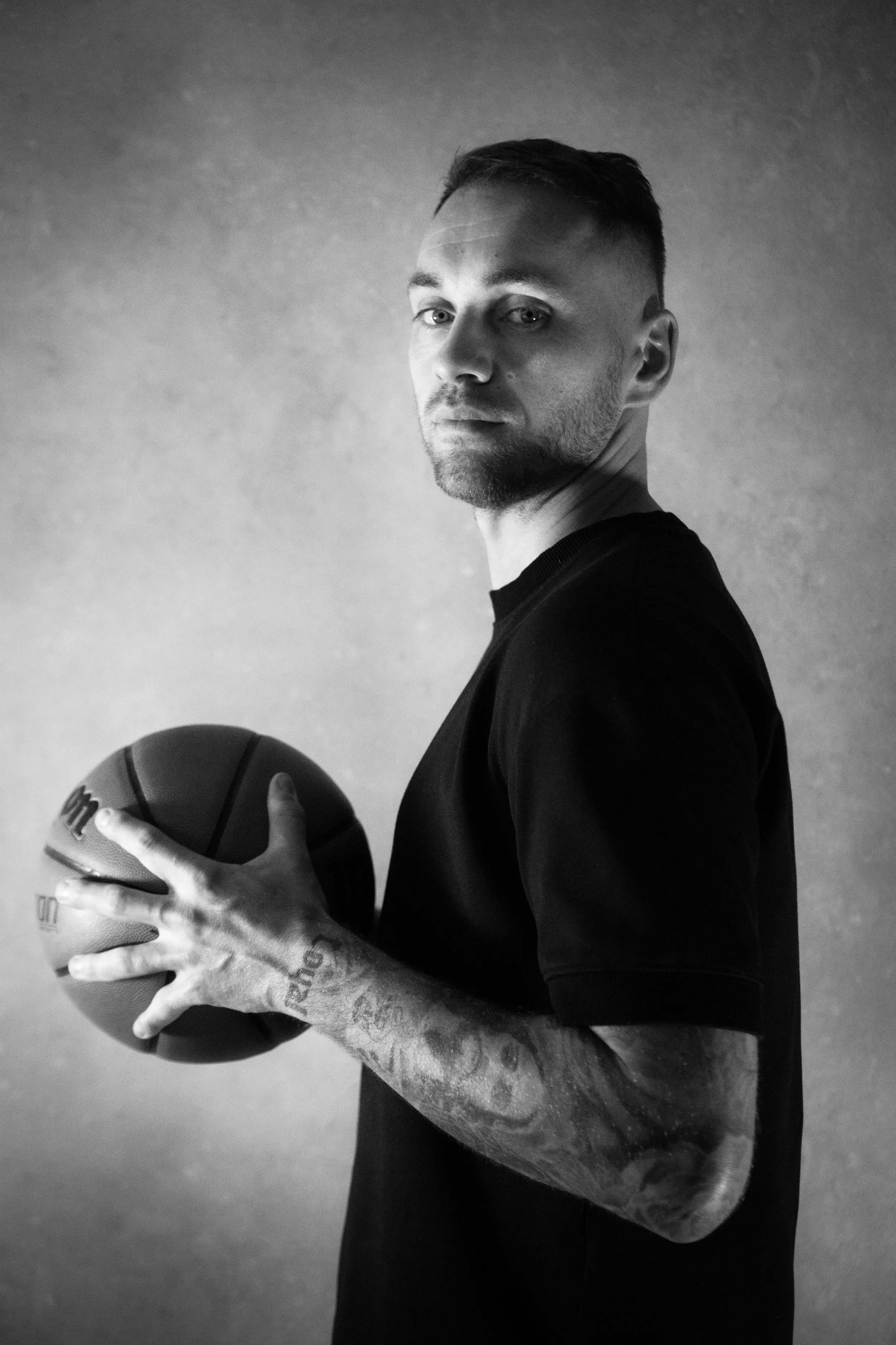 Schwarz-Weiß-Fotografie eines Mannes mit Tattoos, der einen Basketball hält, vor einem neutralen Hintergrund.