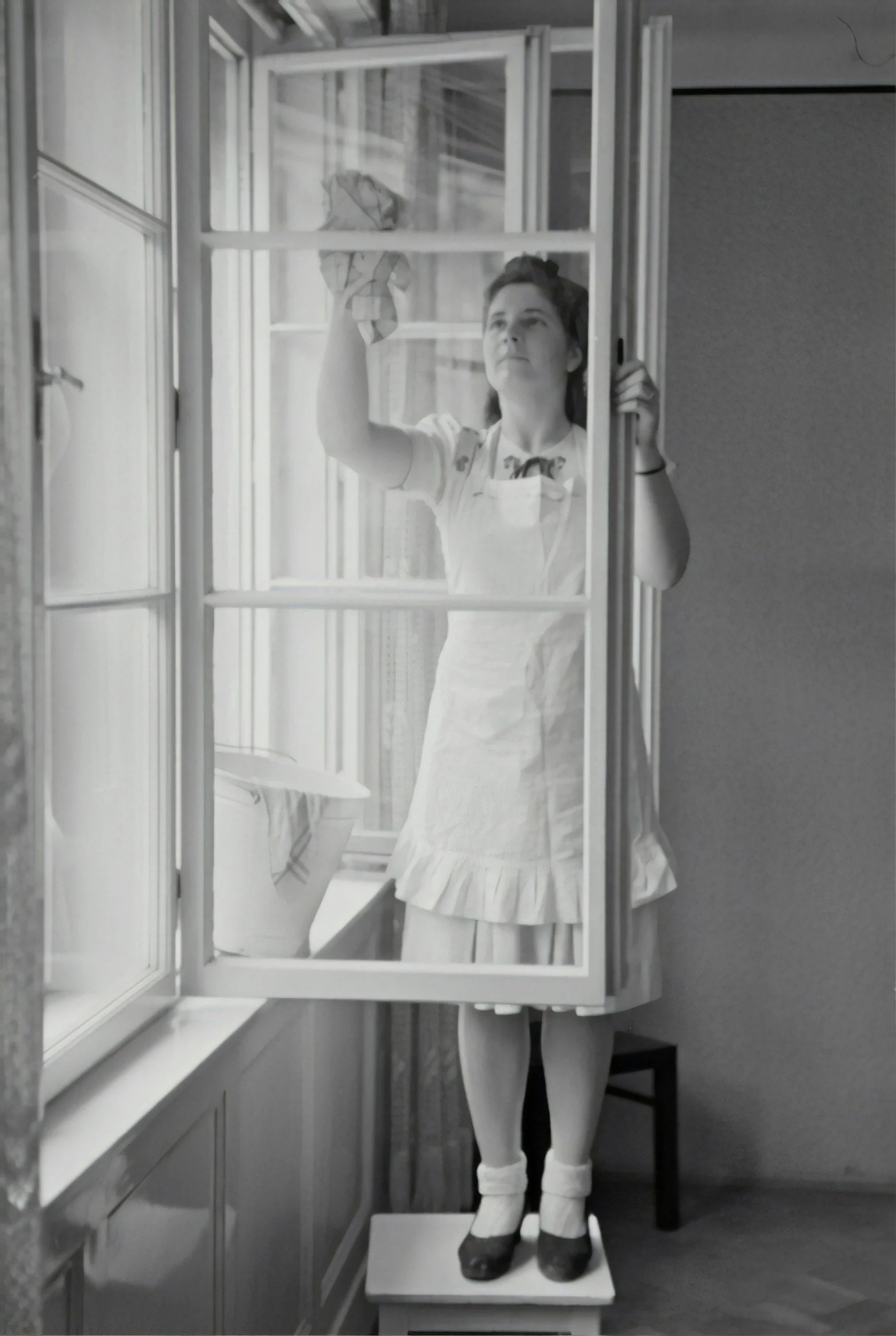 Black and white photo of a young woman standing on a small platform, looking out of a large window. She is reaching up to clean or adjust the window with a cloth. She is wearing a light-colored dress, white apron, and stockings. The room appears to be a vintage-style kitchen with cabinets and a countertop.