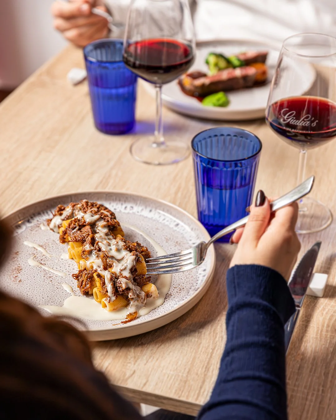 A dining table with a plate of pasta topped with meat and sauce, several glasses of red wine, and blue glasses. A person's hands are visible, one holding a fork with pasta and the other holding a knife.