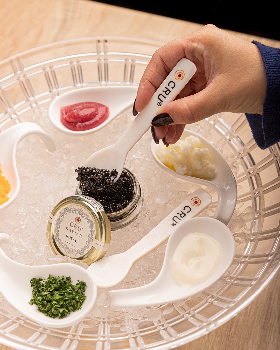 A hand holding a CRU caviar spoon over a glass dish filled with ice and various bowls of condiments and garnishes, including black caviar, chives, lemon cream, and a red sauce.