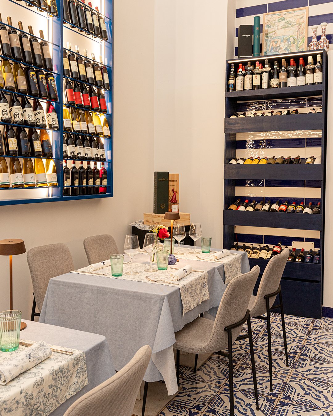 Interior of a restaurant or wine tasting room featuring a table set with wine glasses, napkins, and a small floral centerpiece. In the background, there are large wine racks filled with bottles on the walls, and the floor has decorative blue and white tiles.