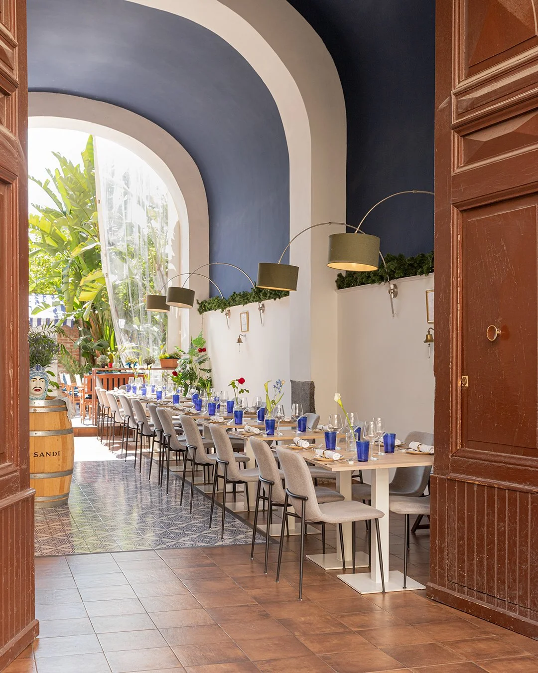 A decorated dining area with tables set for a holiday celebration, featuring Christmas decorations like pine cones, garlands, and red ribbons. The room has blue and white walls, large windows, and modern lighting fixtures.