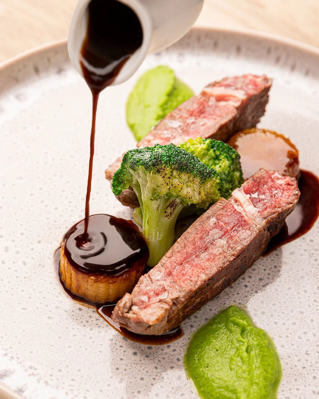 Seared steak slices with broccoli, green sauce, and dark sauce being poured on a plate