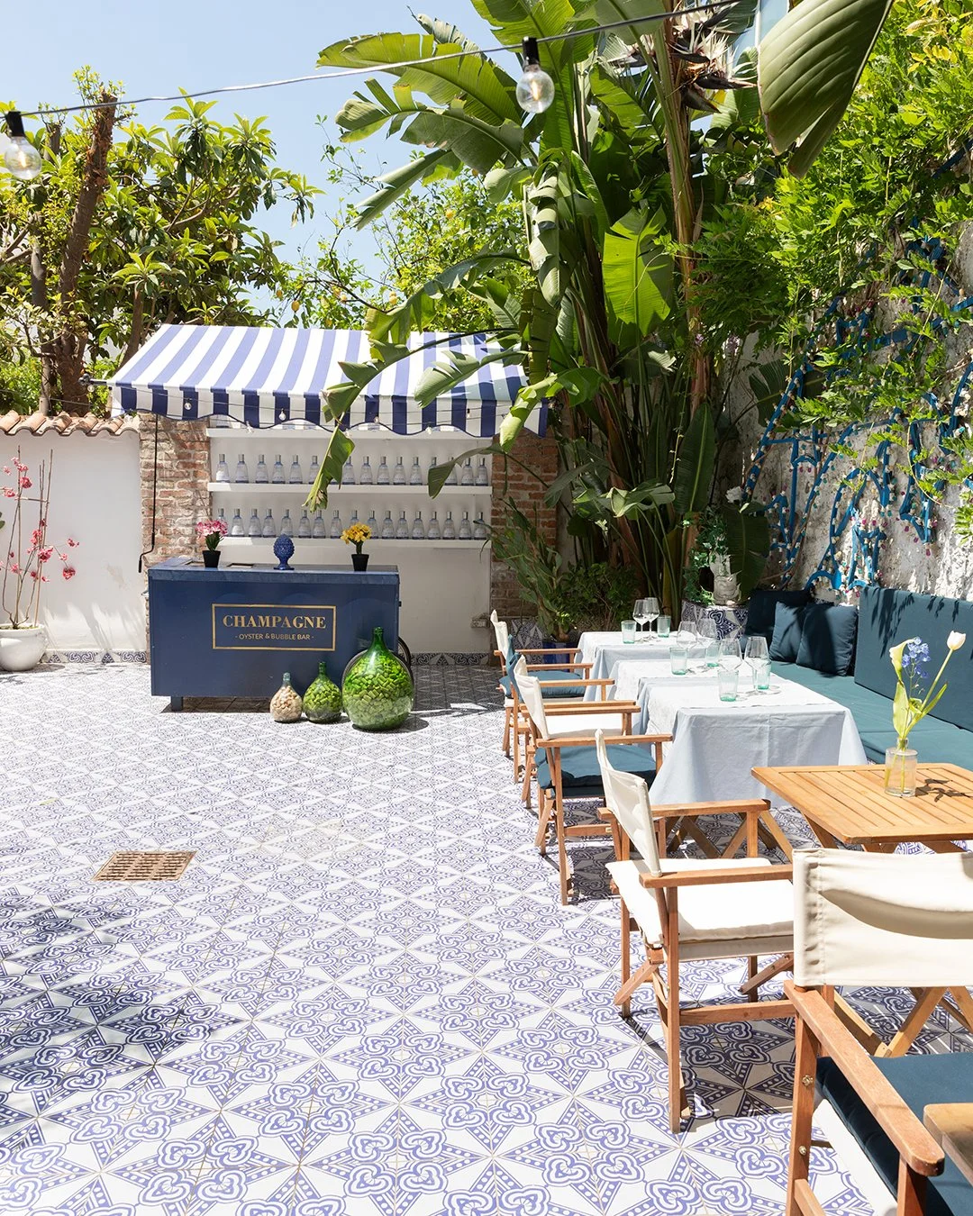 Outdoor dining area with tables set with white tablecloths, glasses, and napkins, surrounded by greenery, including large leaves and trees; a bar with a sign that says "Champagne" in the background; decorative plants and flowers; patterned tile floor.