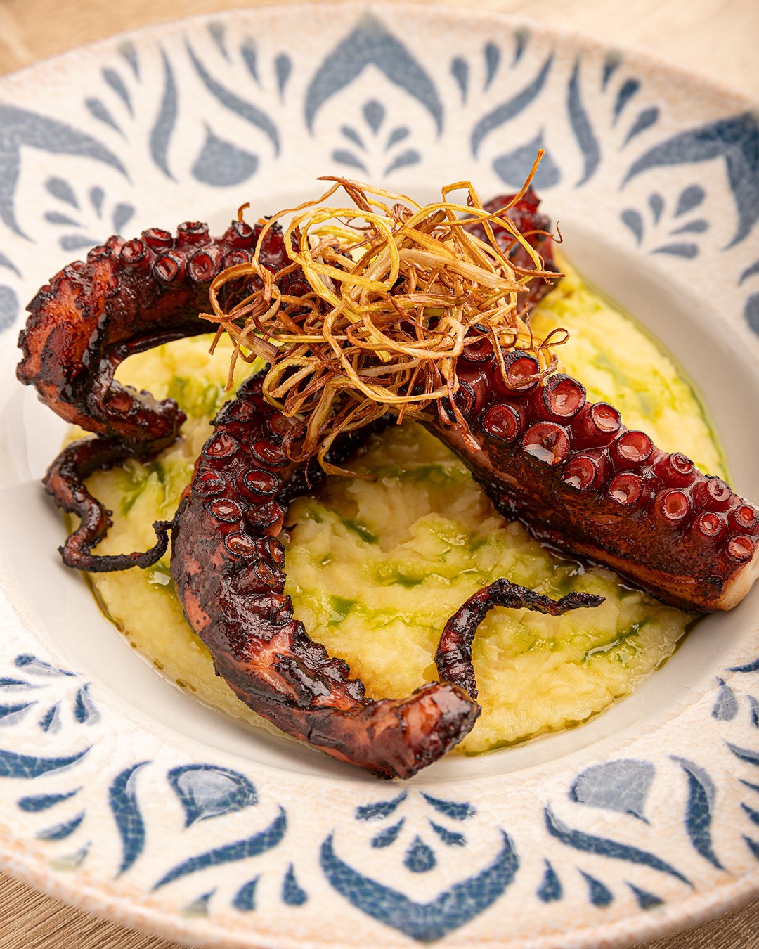 Grilled octopus tentacles served over mashed potatoes with a drizzle of green sauce, topped with crispy fried shredded vegetables, in a decorative blue and white bowl.
