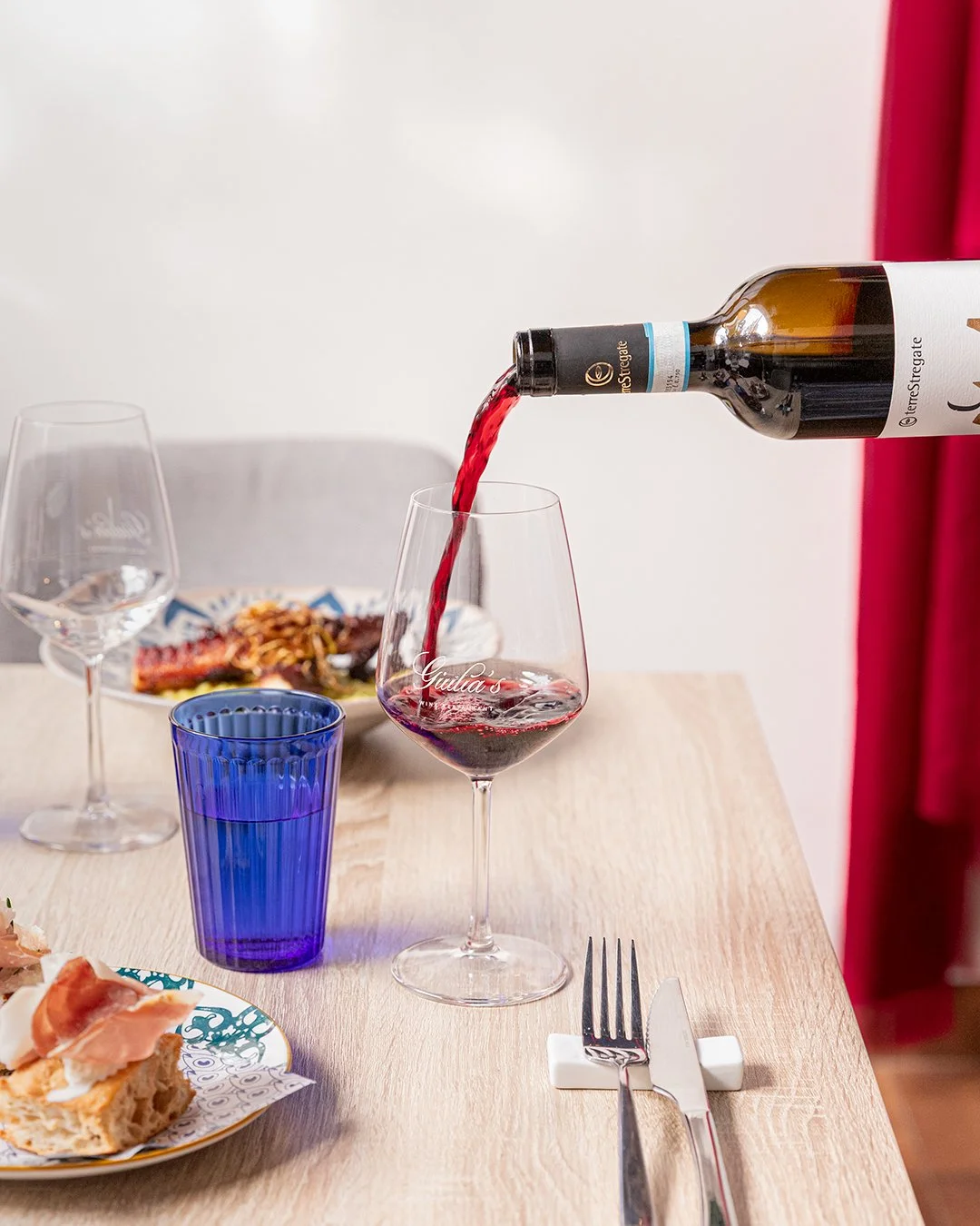 Red wine being poured into a glass on a dining table with a plate of food in the background and additional glasses and utensils on the table.