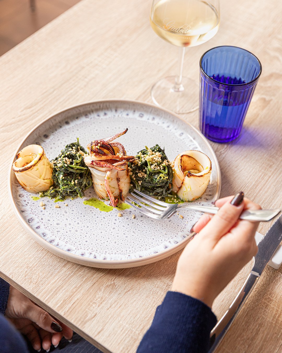 A plate with stuffed squid and greens, two glasses of wine, including a glass of white wine, on a wooden table.