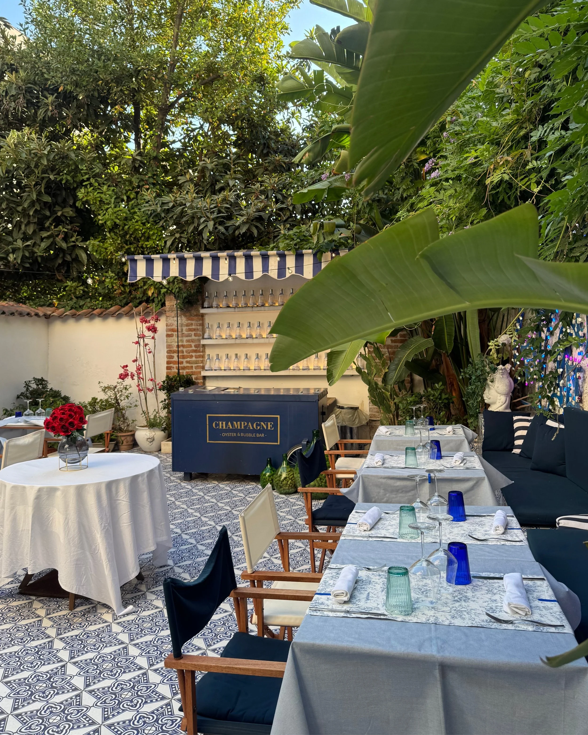 Outdoor dining area with tables set with white tablecloths, glasses, and napkins, surrounded by greenery, including large leaves and trees; a bar with a sign that says "Champagne" in the background; decorative plants and flowers; patterned tile floor.