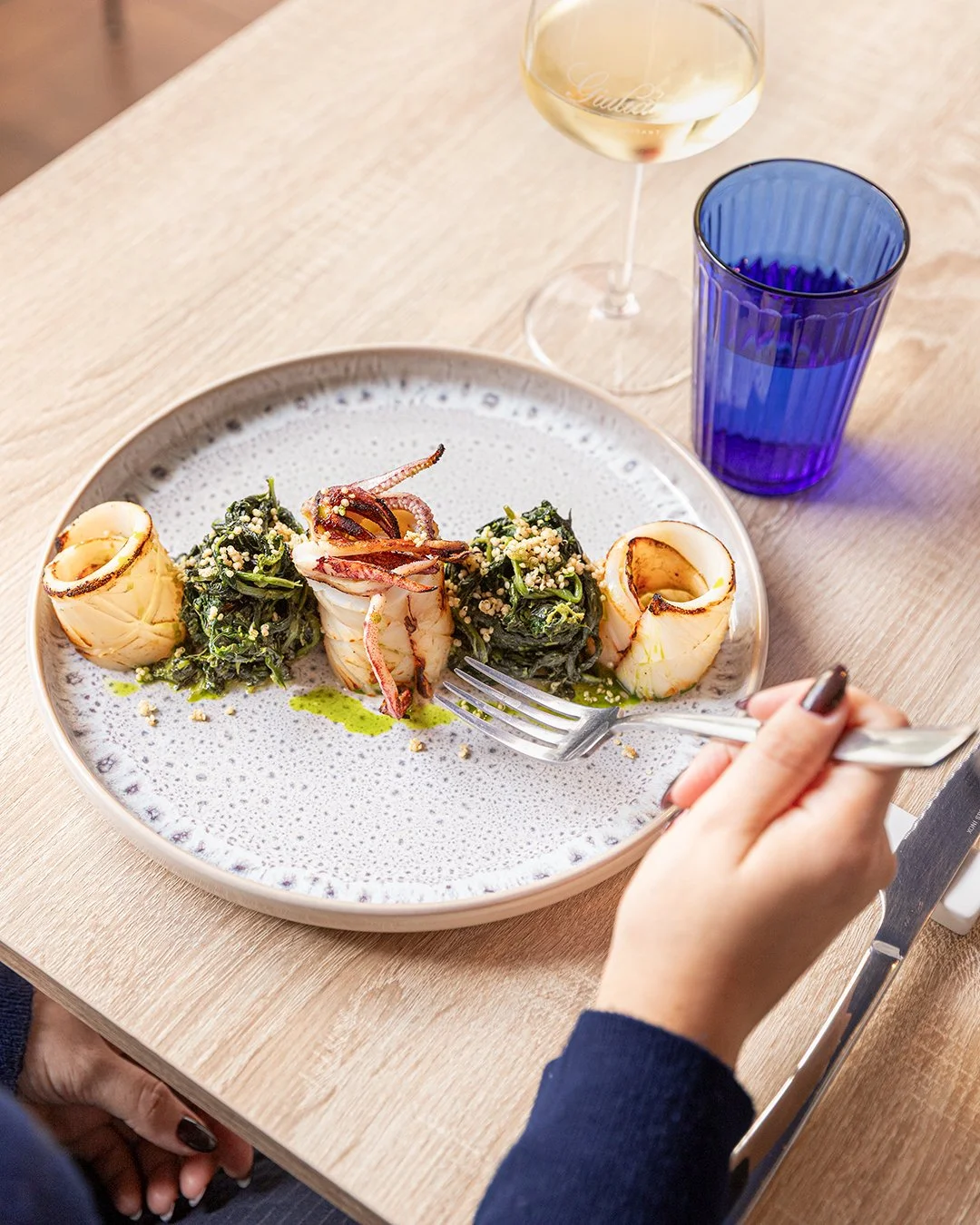 A plate of gourmet seafood with grilled calamari and greens, paired with a glass of white wine and a tumbler of water on a wooden table.
