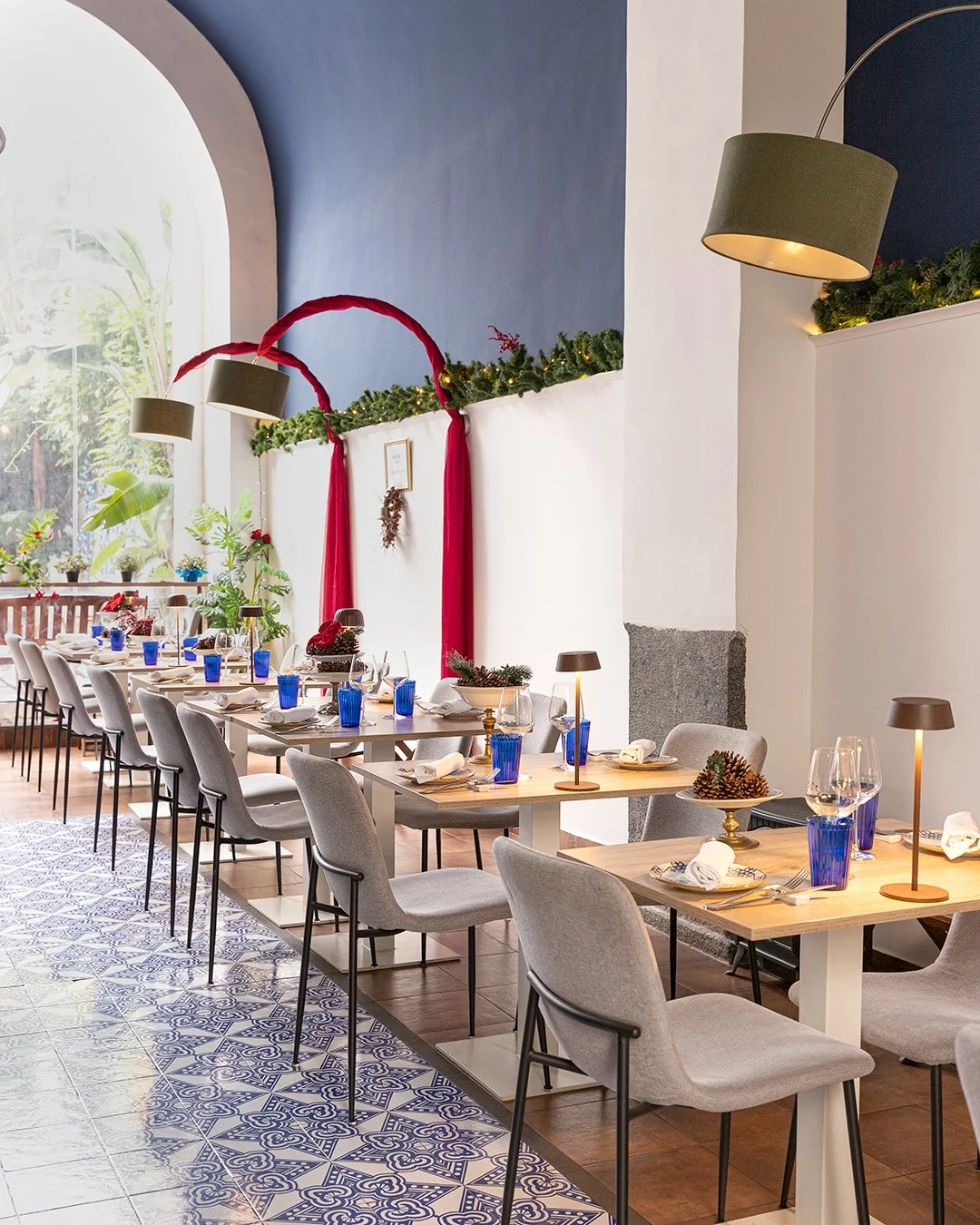 A decorated dining area with tables set for a holiday celebration, featuring Christmas decorations like pine cones, garlands, and red ribbons. The room has blue and white walls, large windows, and modern lighting fixtures.