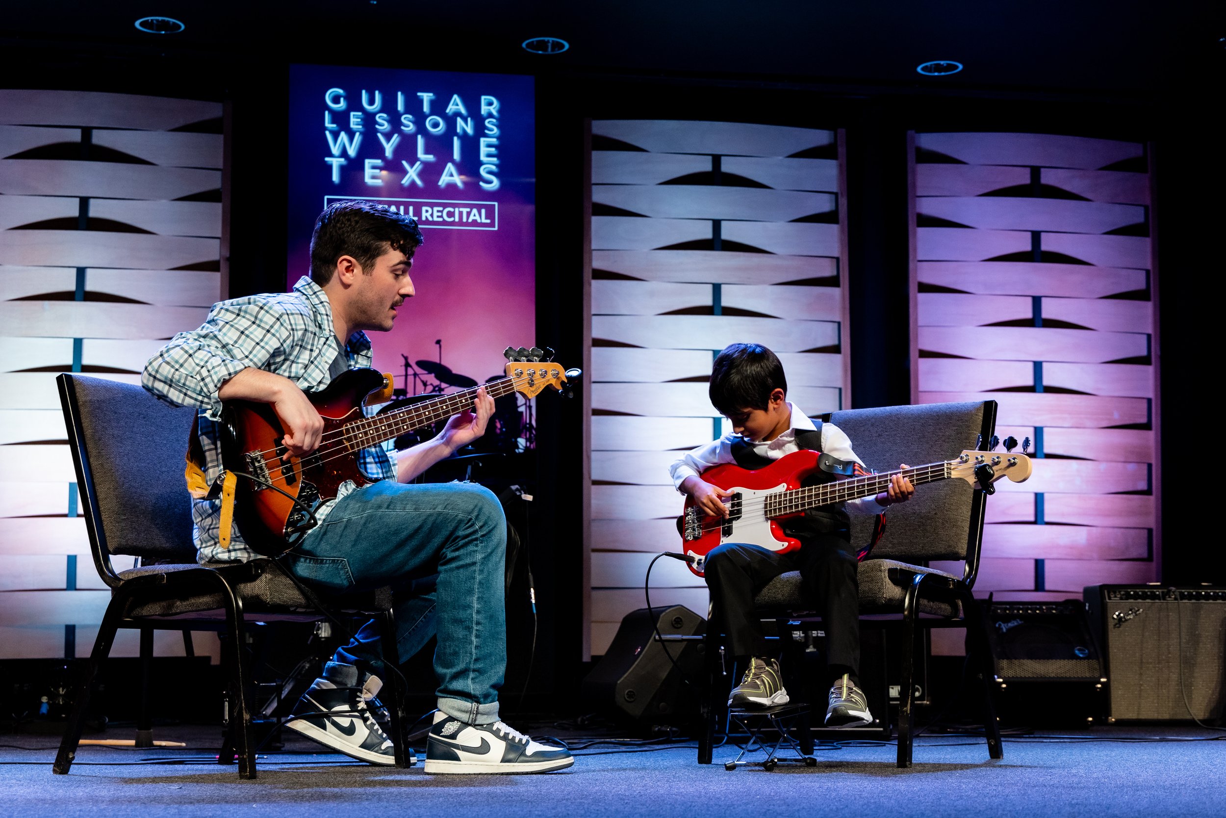 Ian teaches bass, guitar, and ukulele at the Wylie School of Music. The photo shows him accompanying one of his students, a young boy, who is playing the bass guitar at his music recital in Wylie, TX.