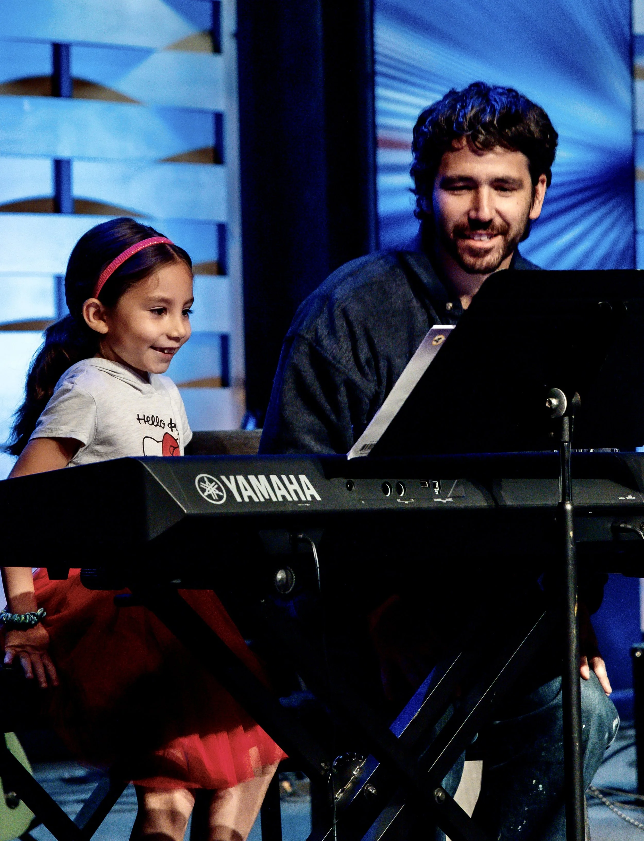 Spencer K. teaches piano, guitar, and bass at the Wylie School of Music. Image shows Spencer with one of his piano students, smiling at her piano recital in Wylie.