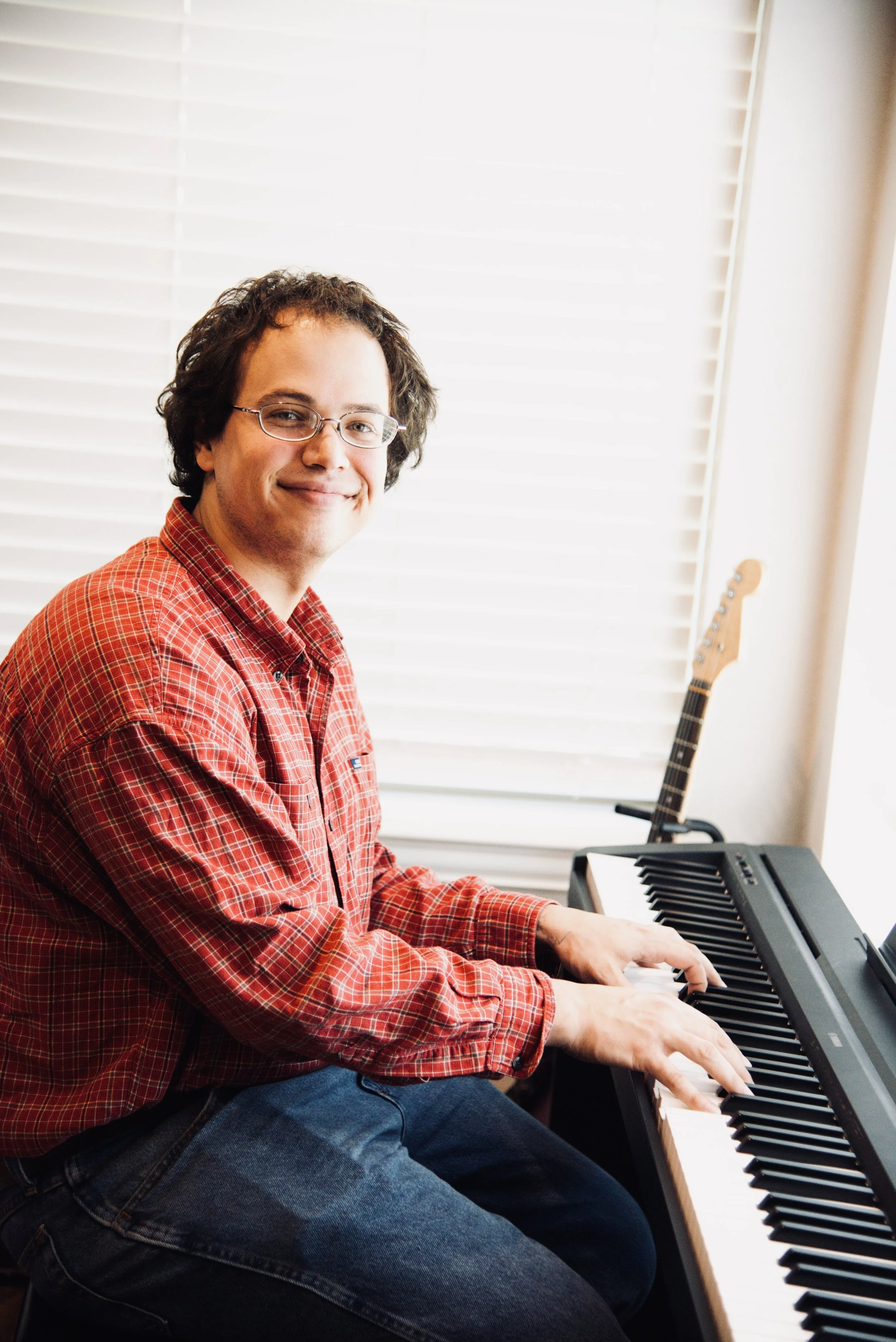 Joseph M. teaches piano, guitar, and bass at the Wylie School of Music. Photo shows him smiling at his keyboard with a guitar in the background.