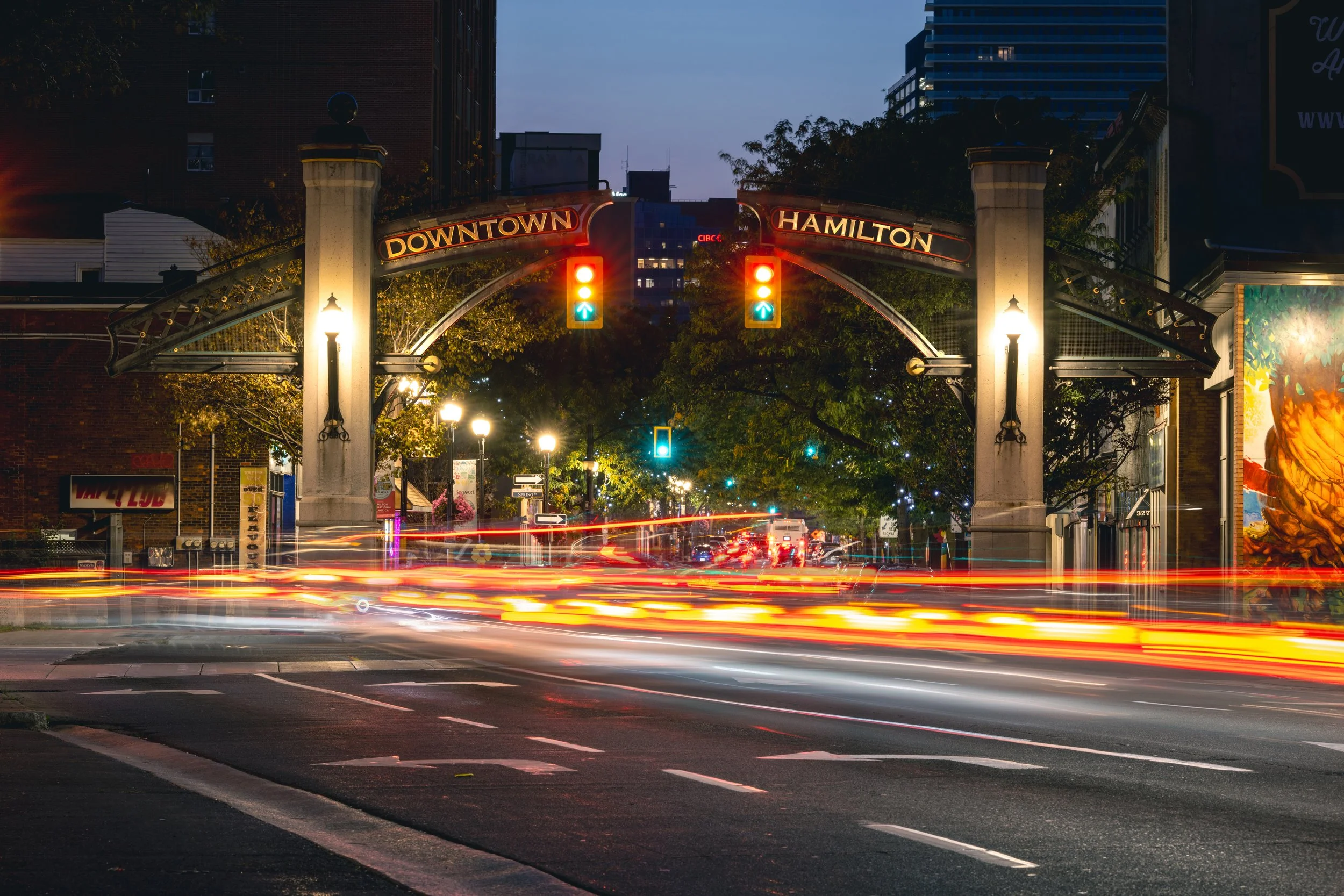Nighttime view of the entrance to downtown Hamilton with illuminated signs, traffic lights, and light streaks from moving vehicles on the street.