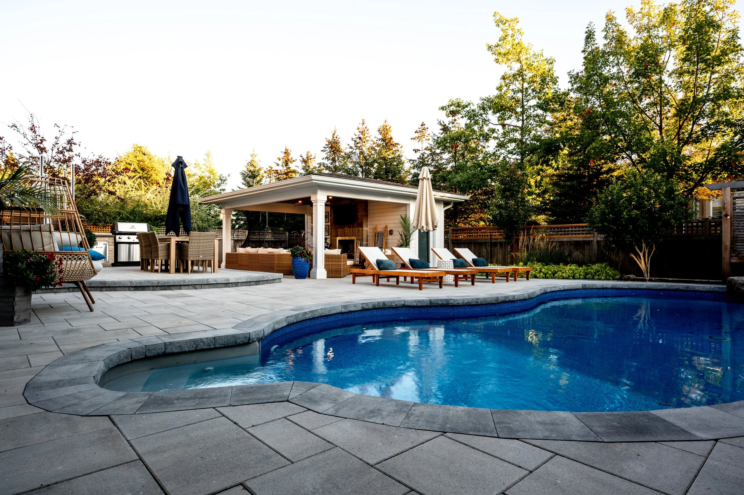 Backyard with swimming pool, outdoor seating, lounge chairs, and a covered patio area surrounded by trees