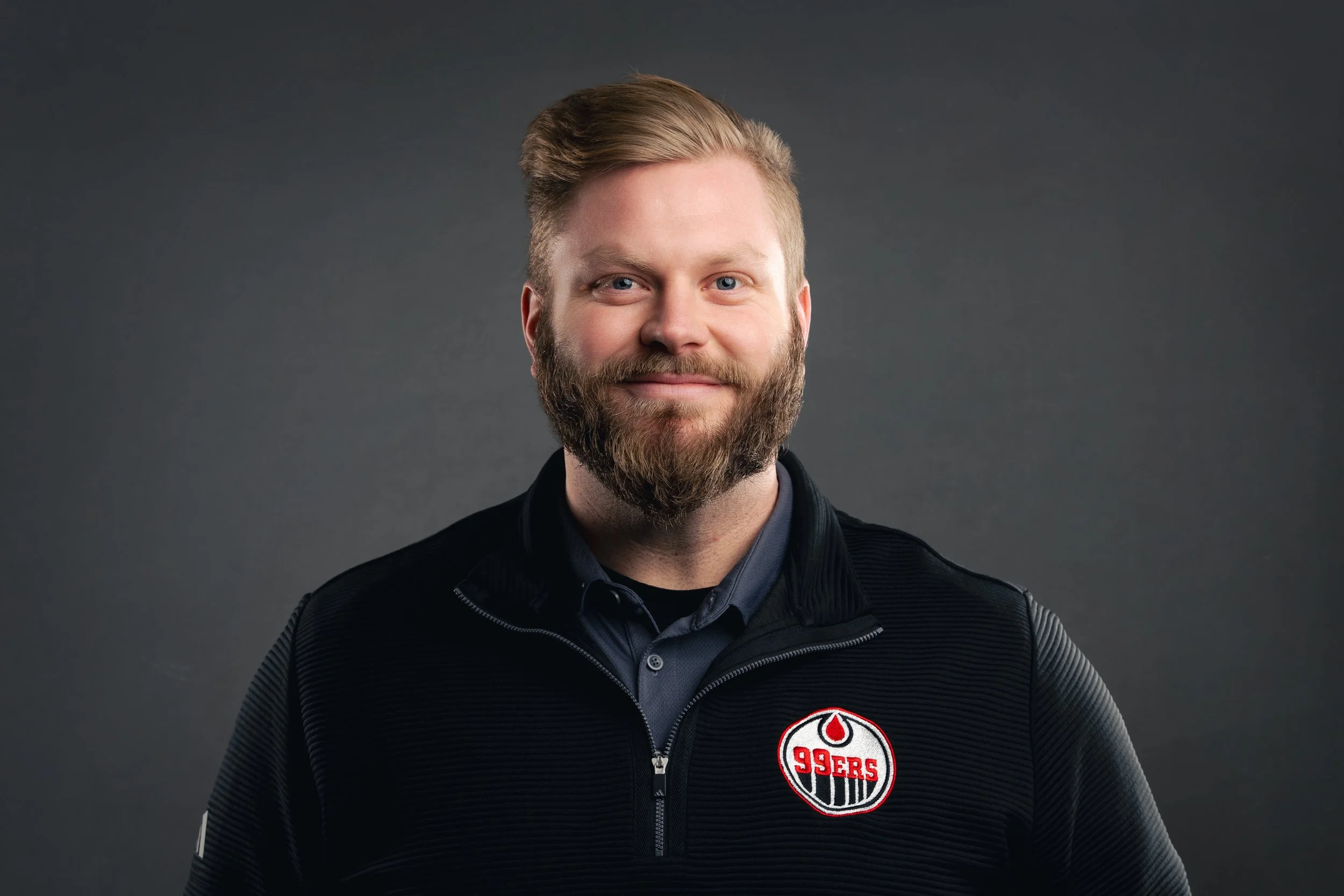 A smiling man with a beard and blonde hair, wearing a black jacket with the San Francisco 49ers logo on it, against a gray background.