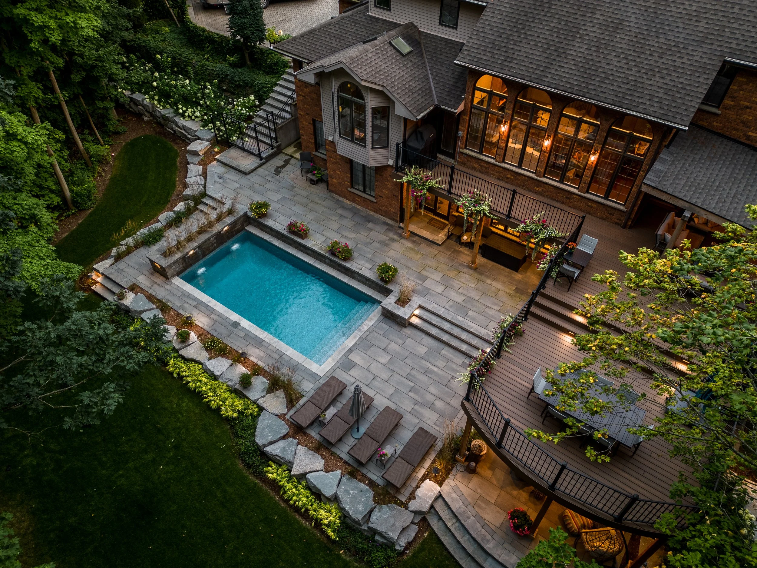 Aerial view of a backyard with a swimming pool, patio seating, steps, landscaped garden, and a house with large windows and multiple decks surrounded by trees and greenery in the evening.