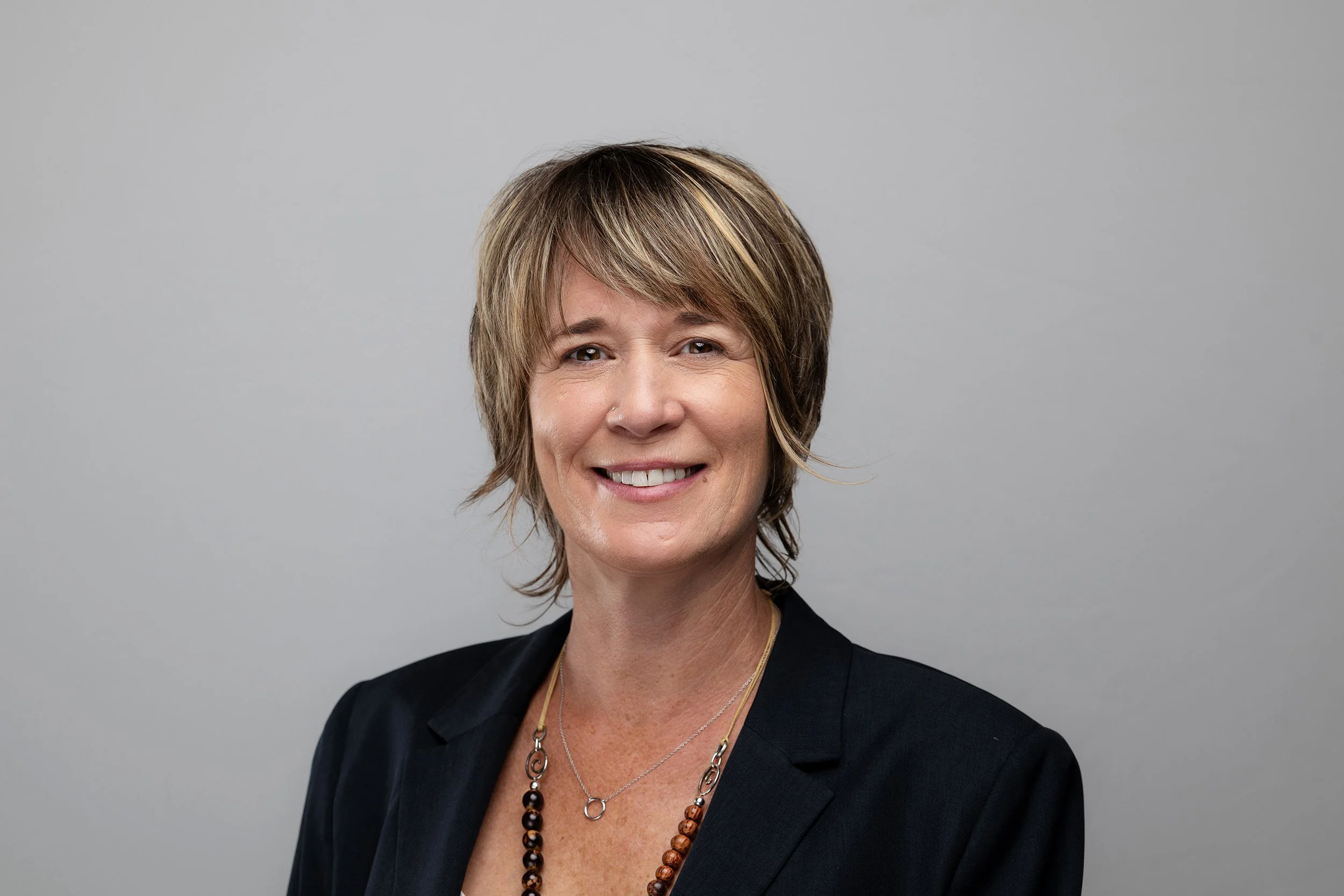 A middle-aged woman with short, layered brown hair with blonde highlights, wearing a black blazer and layered necklaces, smiling against a plain gray background.