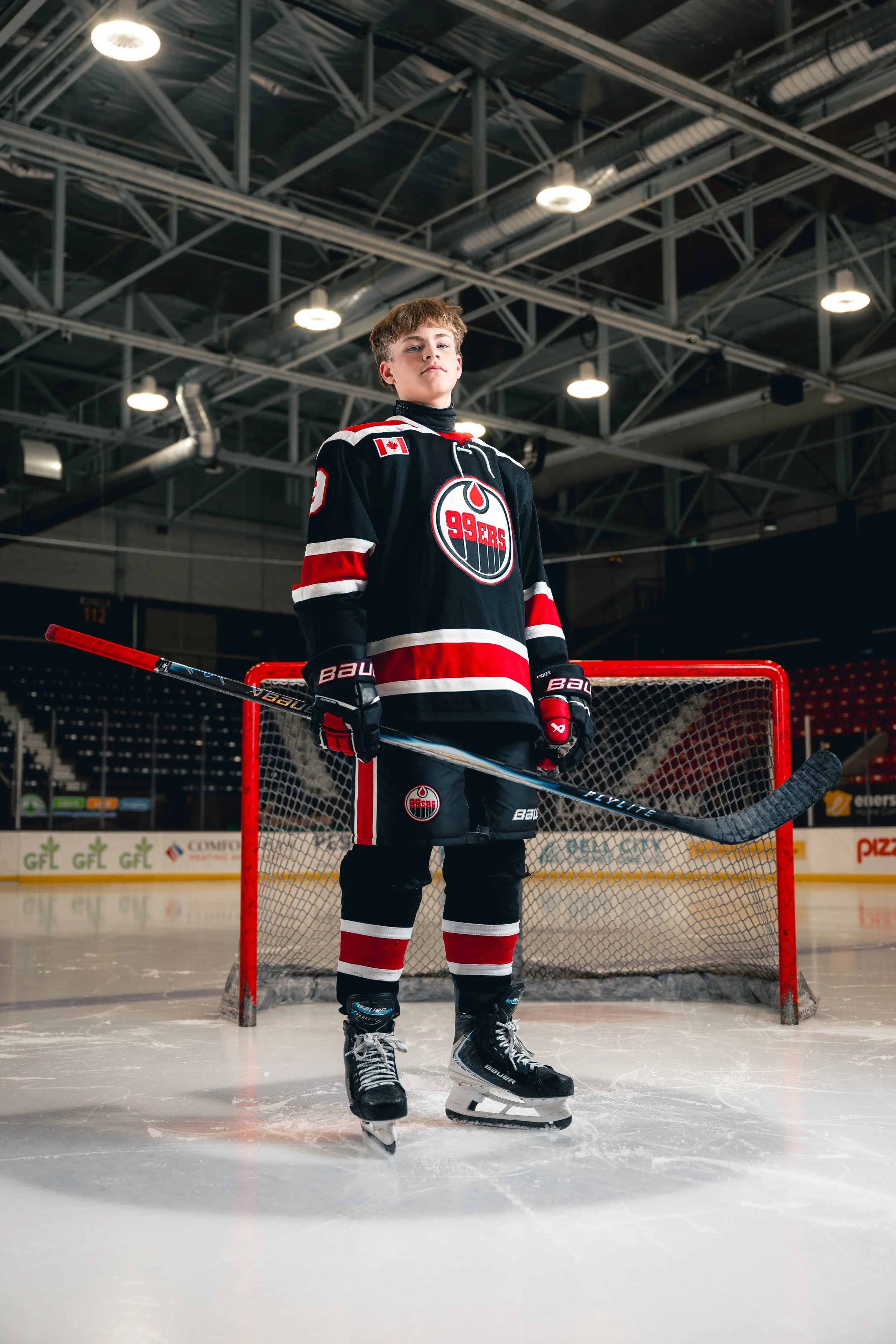 Young male ice hockey player standing in front of goal on ice rink, wearing black, red, and white hockey uniform with number 92, holding hockey stick.