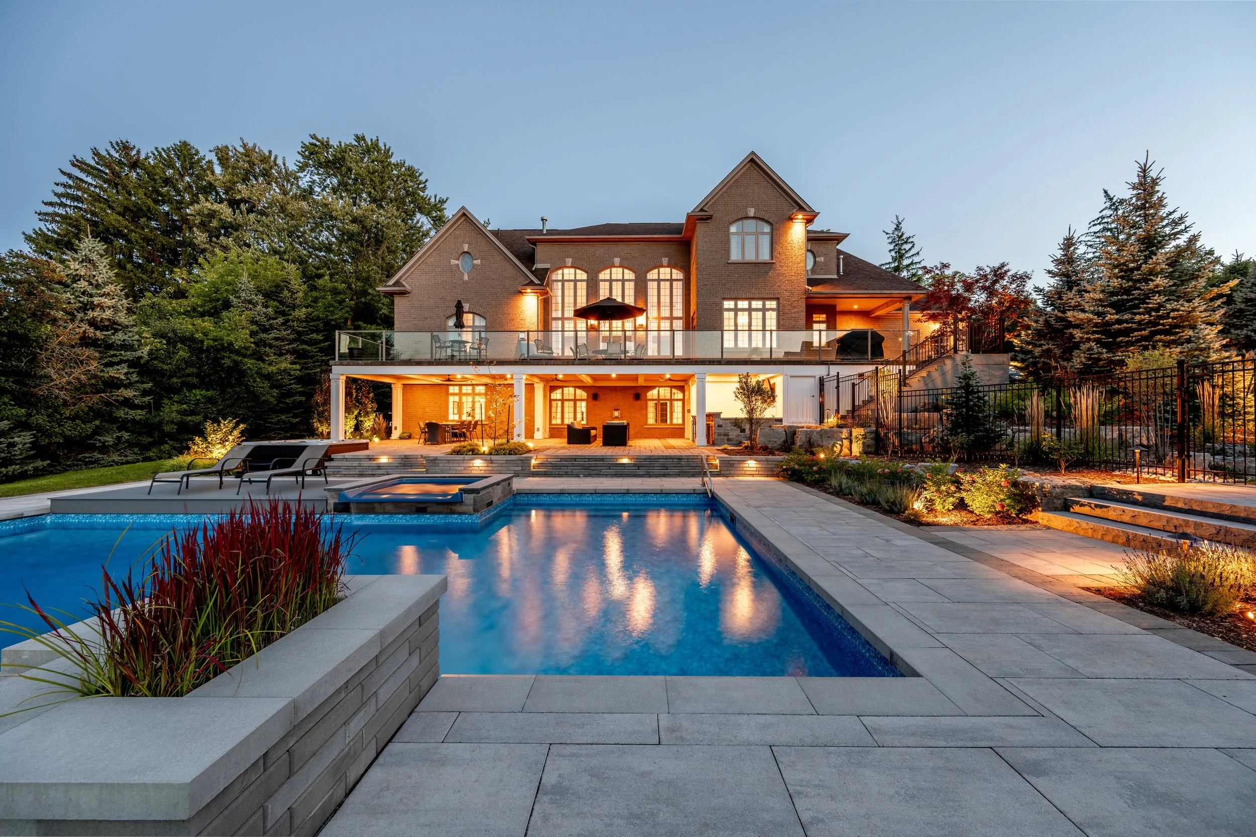 A large brick house with multiple stories, illuminated windows, and a spacious backyard featuring a swimming pool, poolside lounge chairs, and landscaped gardens seen at dusk.