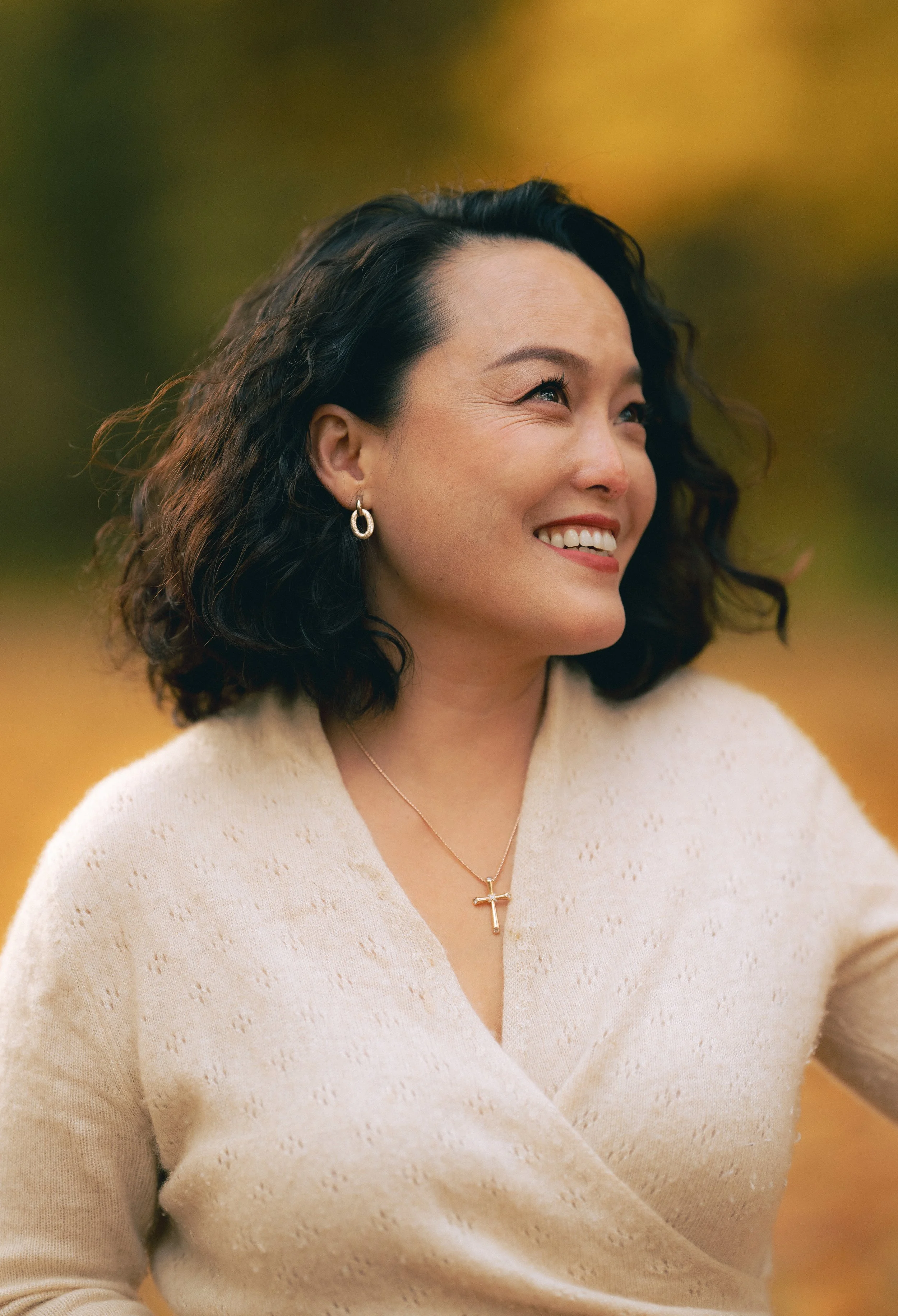 A woman with shoulder-length dark curly hair, smiling and looking to her left, wearing a cream-colored shawl, gold hoop earrings, and a gold necklace with a cross pendant, outdoors during autumn.