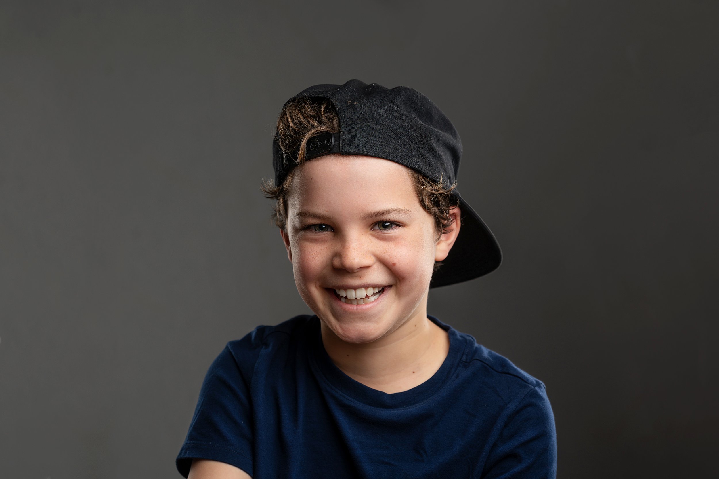 Smiling boy wearing a backward black baseball cap and a navy blue t-shirt, against a gray background.