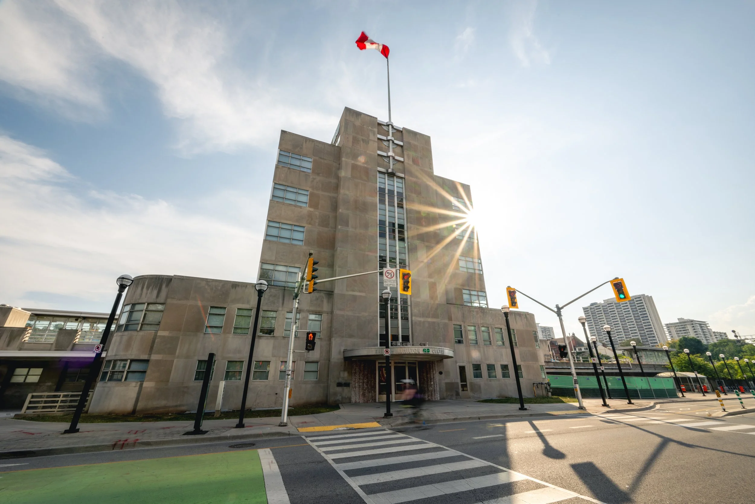 A multi-story concrete building with a Canadian flag on top, illuminated by sunlight, with traffic lights and crosswalks in the foreground and a blue sky with some clouds.
