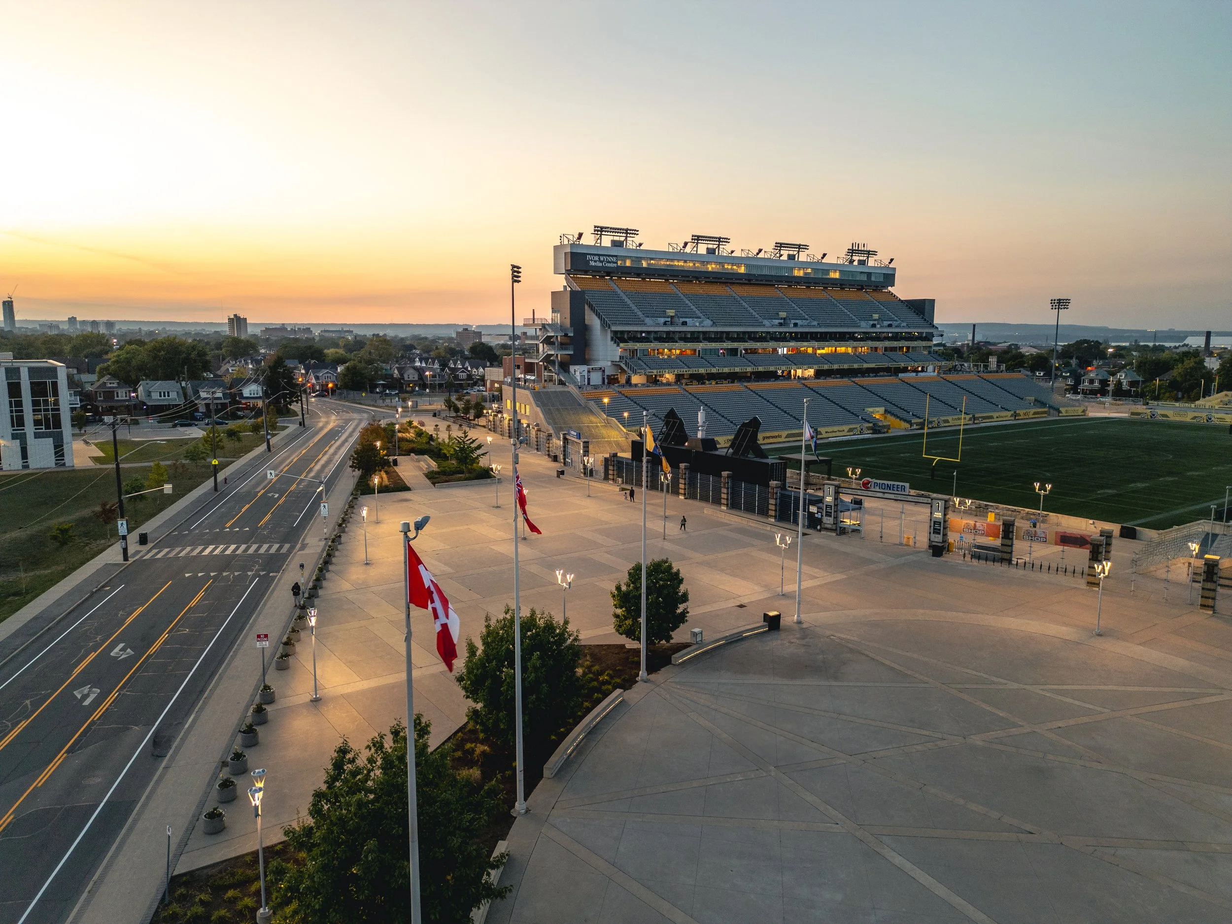 An empty football stadium at sunset with flags flying, light posts on the field, and a quiet surrounding cityscape in the background.