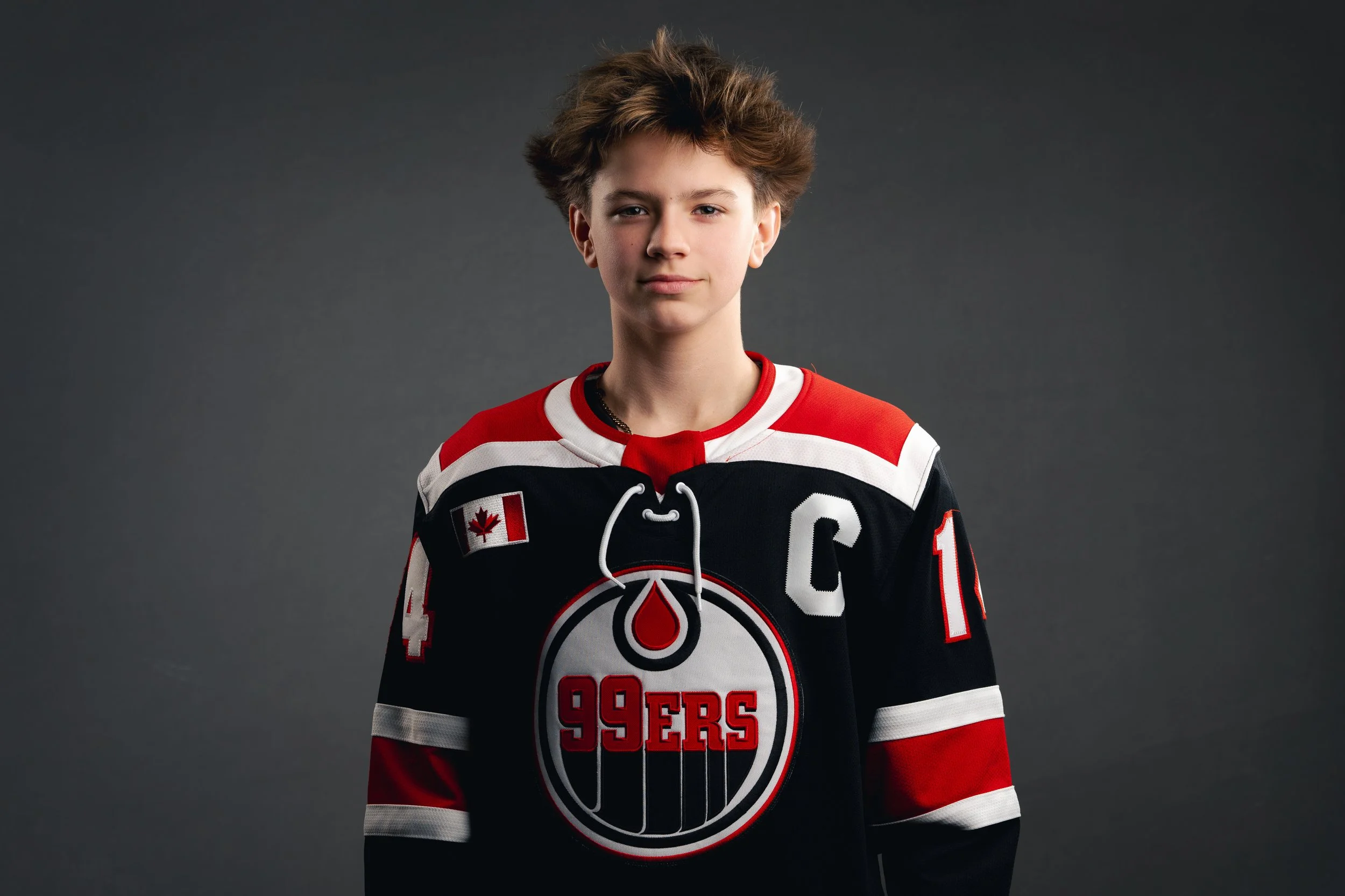 A young boy with messy brown hair wearing a San Jose Sharks hockey jersey standing against a dark gray background.