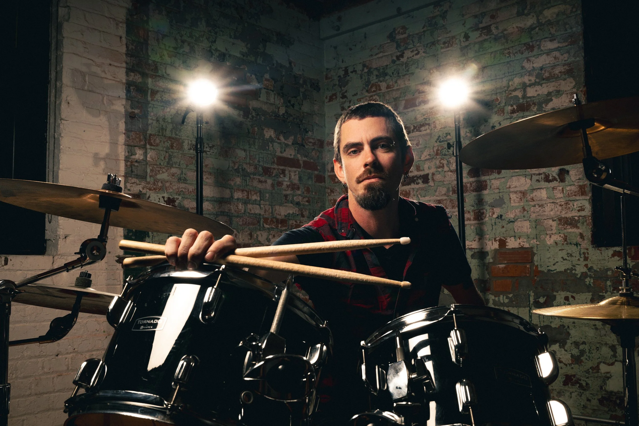A man with short dark hair, facial hair, and a serious expression sitting behind a black drum set in a music studio with brick walls and two bright studio lights.
