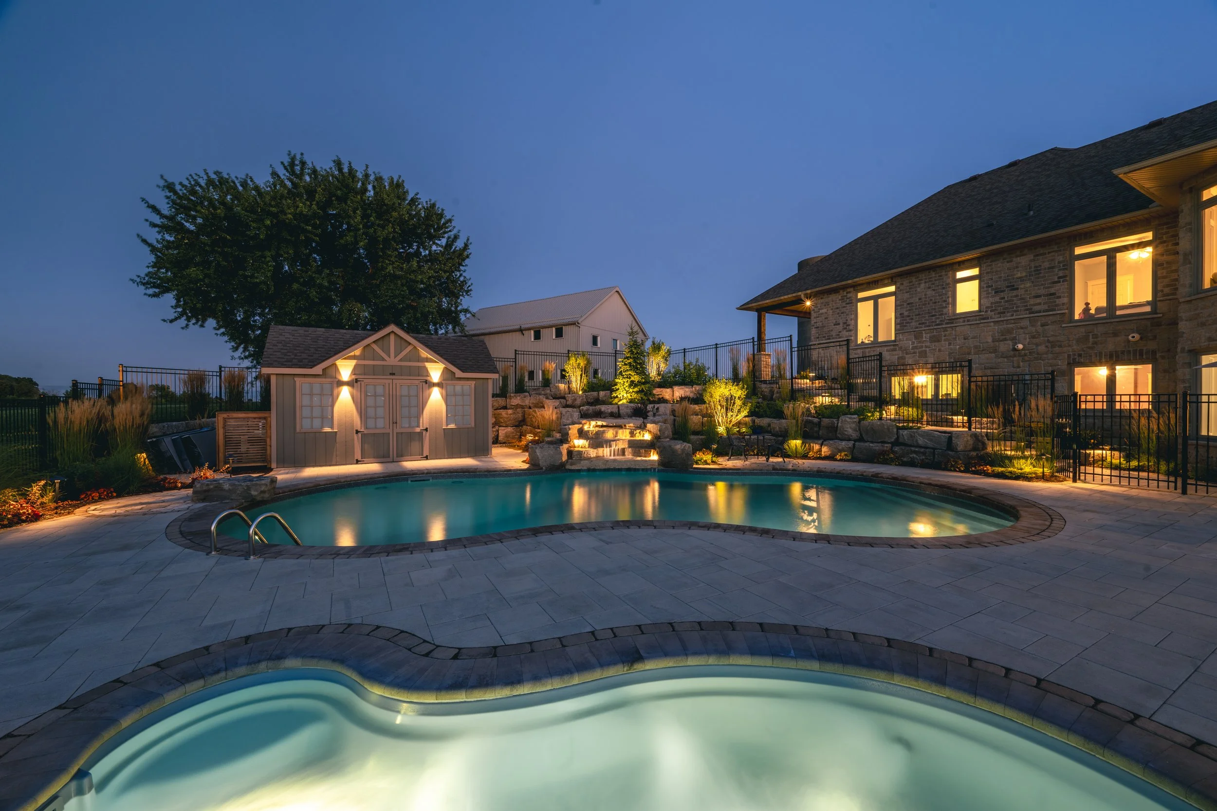 A backyard at dusk featuring a swimming pool with illuminated hot tub in the foreground, a small shed with exterior lighting, a rock waterfall, and a large house with multiple windows and outdoor lighting. Foliage and a large tree are in the backgrou