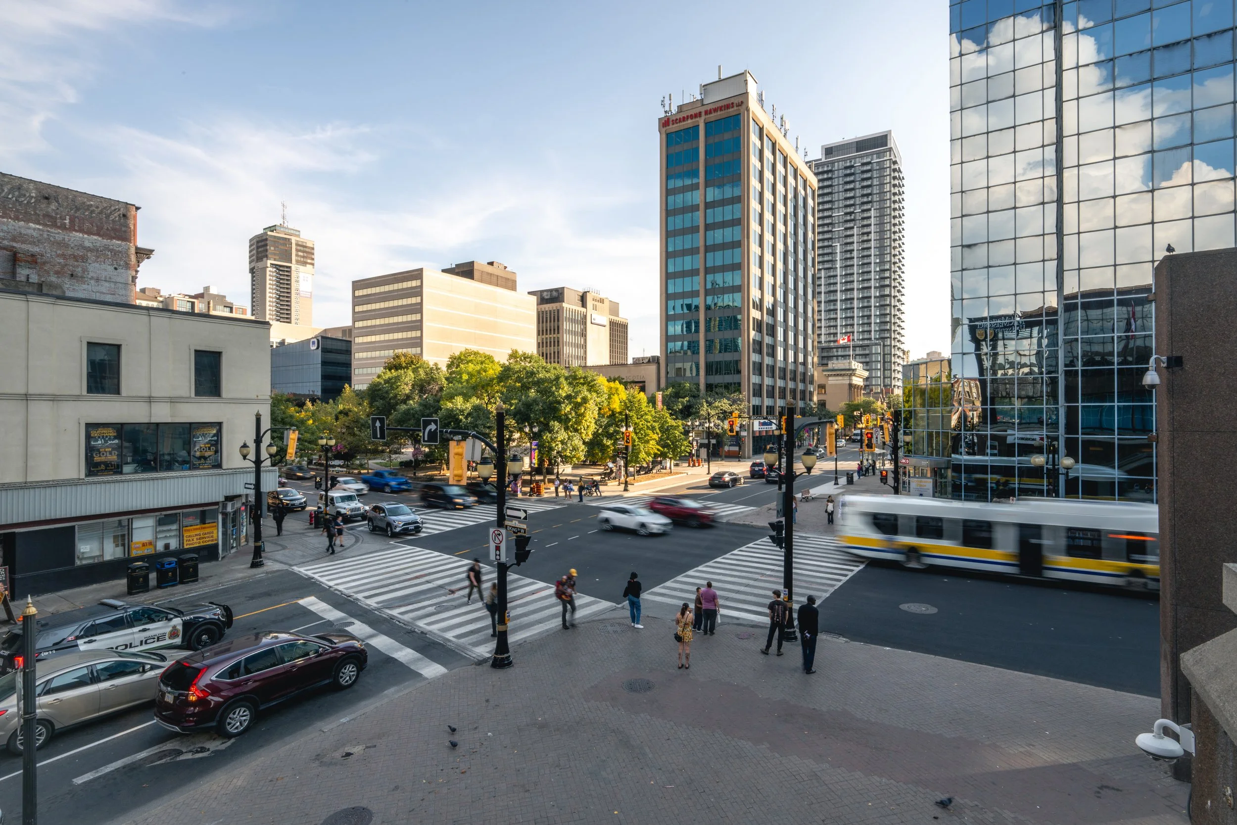 An urban city street scene with crosswalks and cars, a bus in motion, pedestrians crossing, and tall buildings with glass windows reflecting the sky.