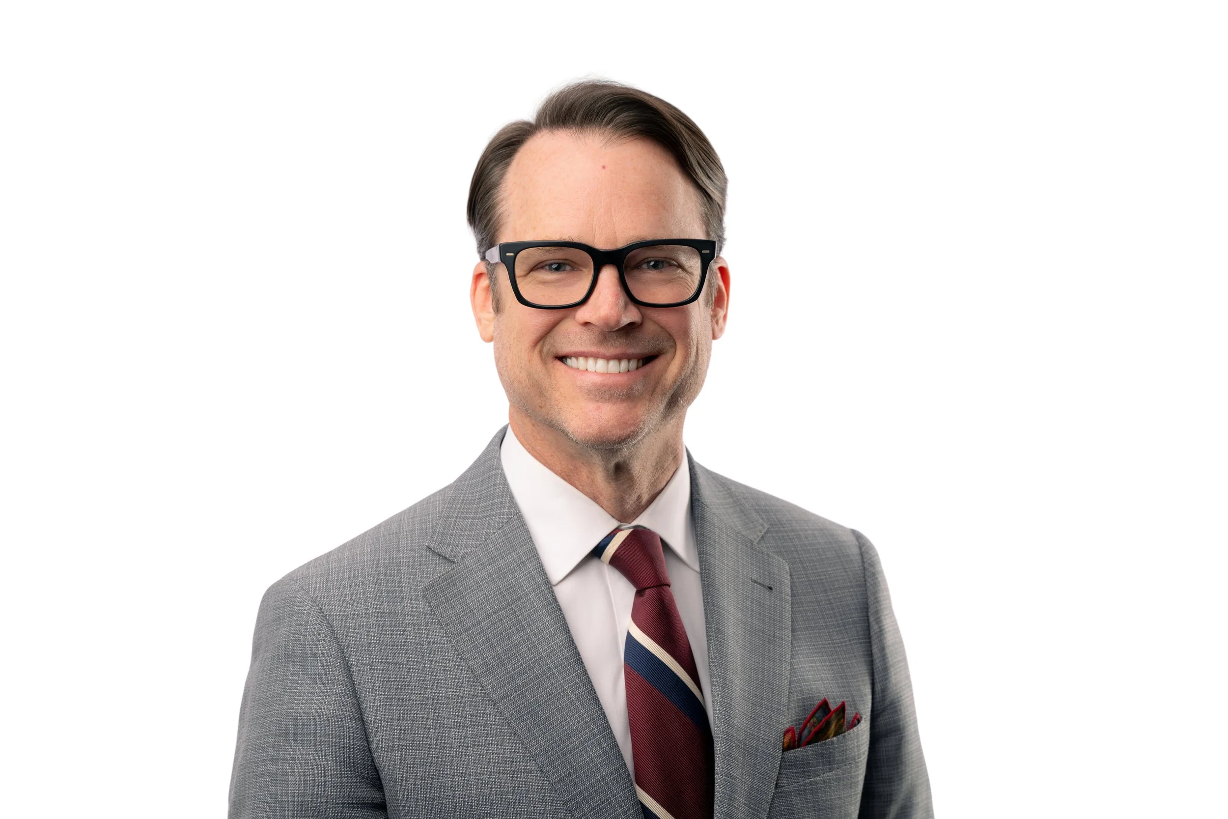 A smiling man wearing glasses, a gray suit, white dress shirt, maroon and navy striped tie, and a pocket square, against a plain white background.