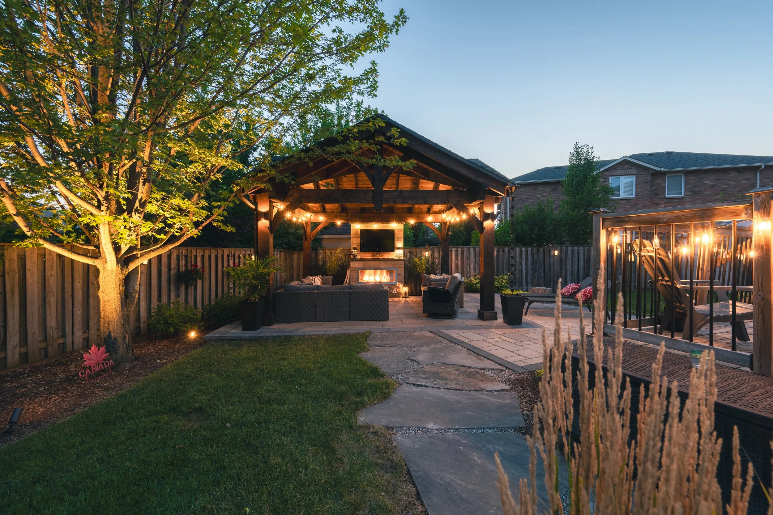 Backyard patio with a wooden gazebo, string lights, outdoor furniture, a fireplace, and a wooden fence, during evening.