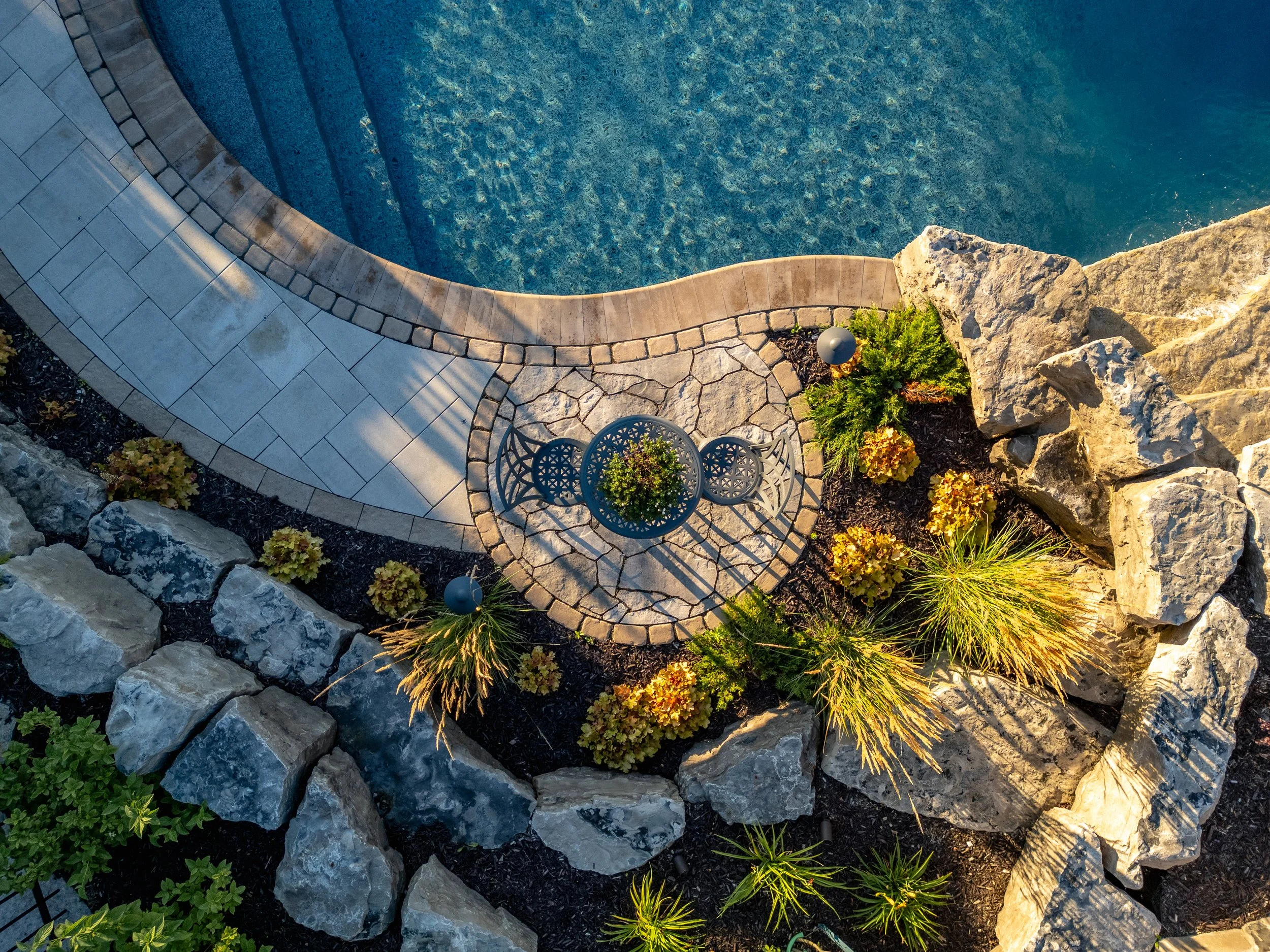 Top-down view of a landscaped garden border with a curved swimming pool, a small round table with two chairs, and various plants and rocks.