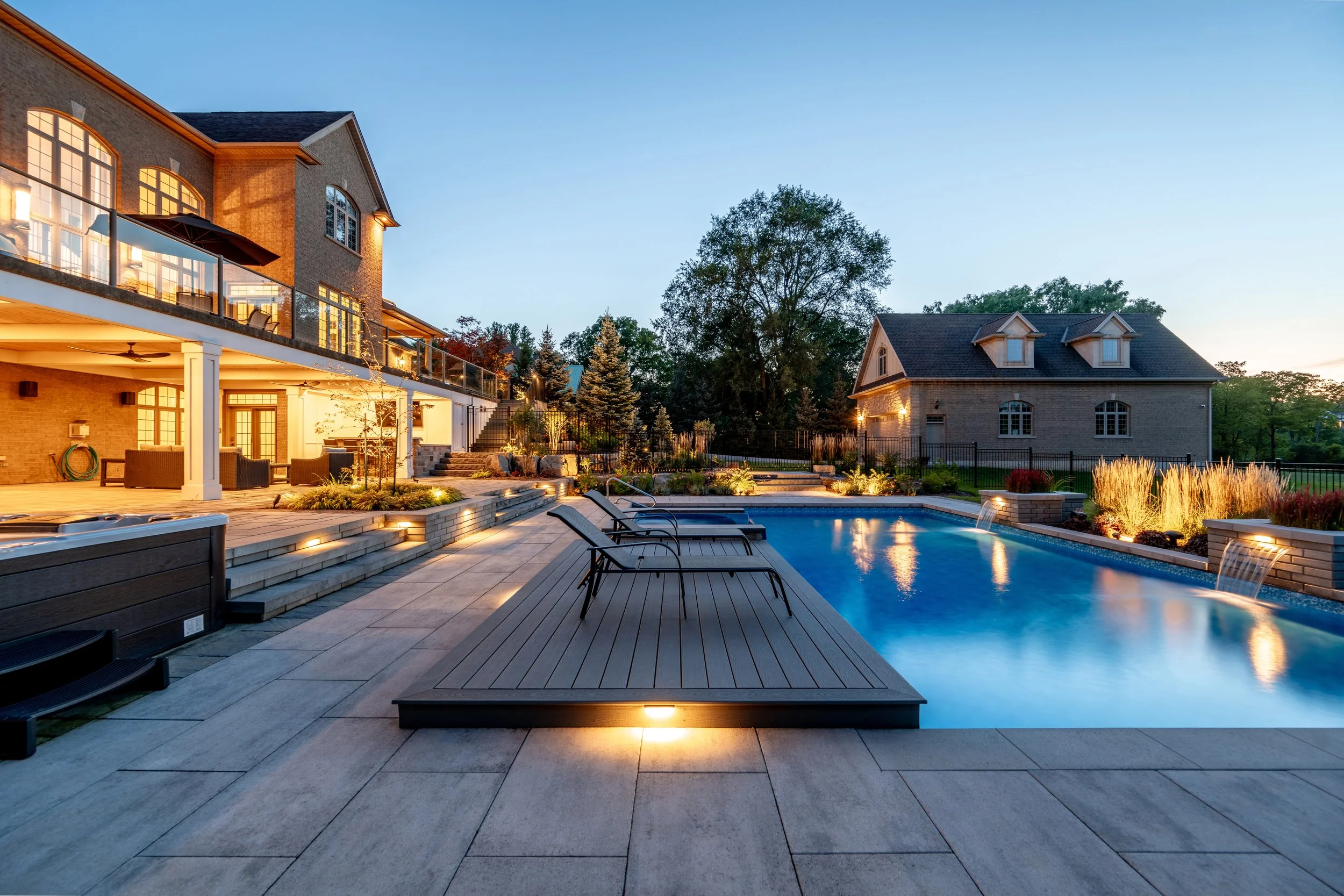 Luxury backyard with a swimming pool, outdoor lounge area, landscaped garden, multi-story brick house with lit windows, and a smaller house in the background during evening.