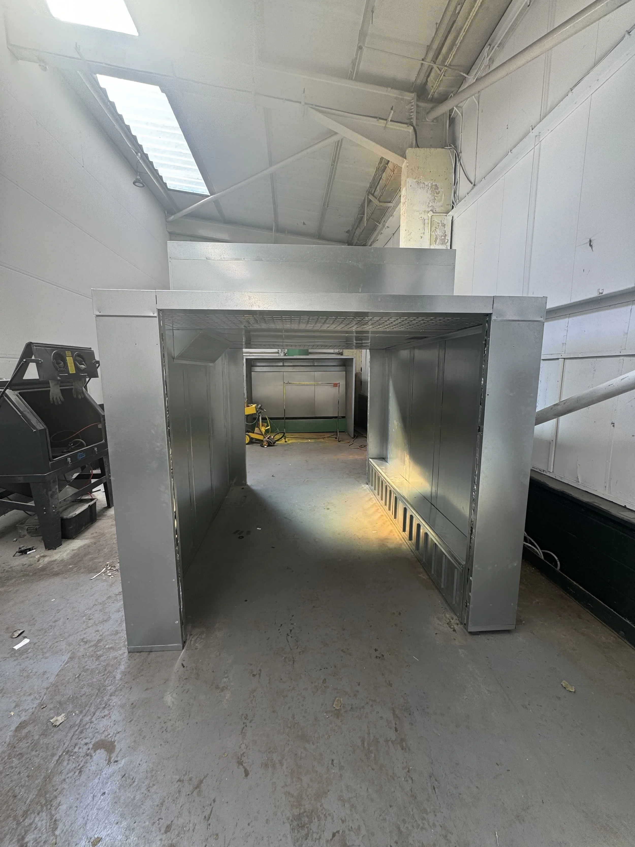 A metal structure oven construction inside a building, with exposed pipes and ceiling, and construction tools visible on the floor.