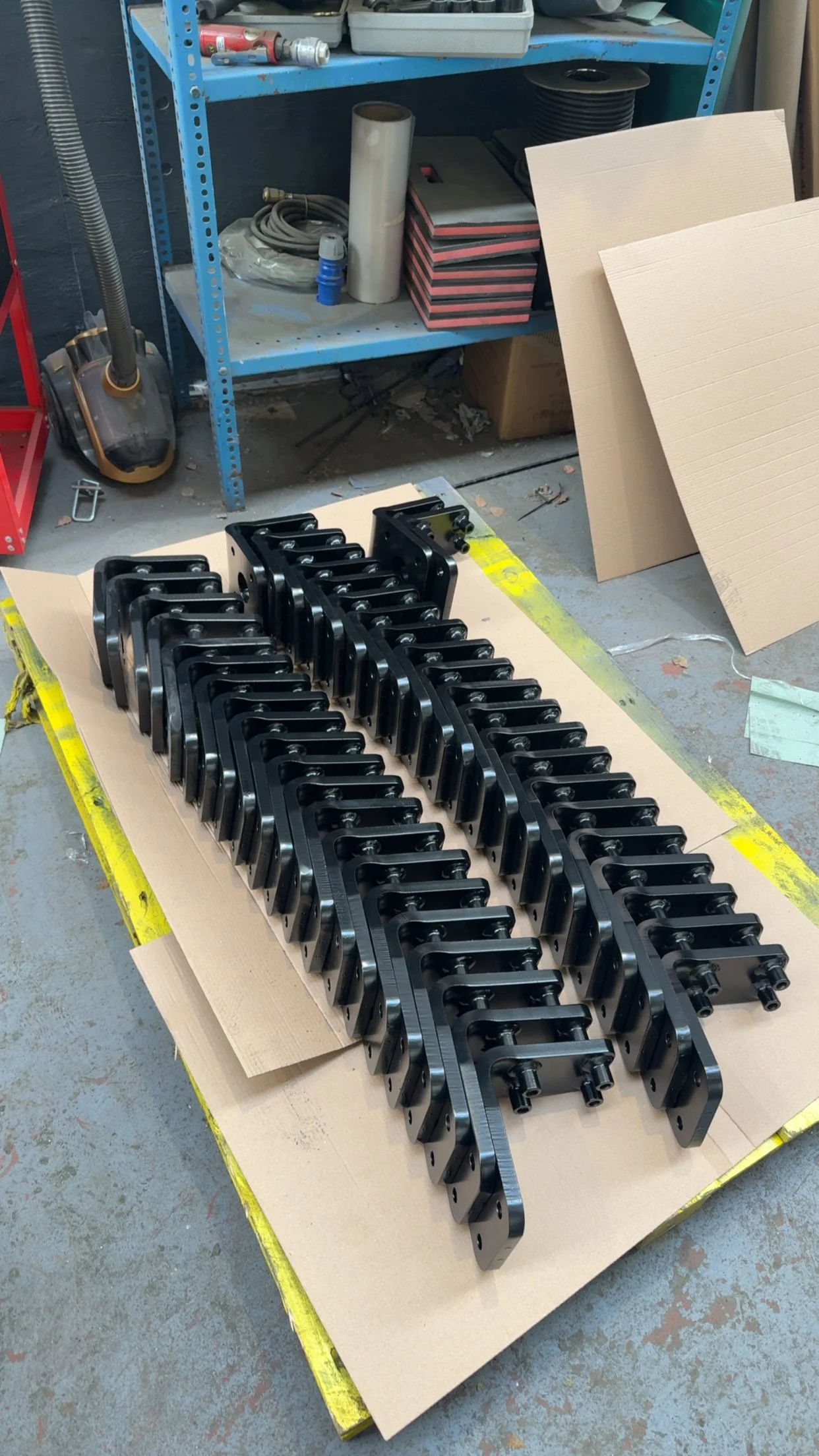 Multiple black metal mechanical parts arranged on a large piece of cardboard in a workshop, with shelves and tools in the background.