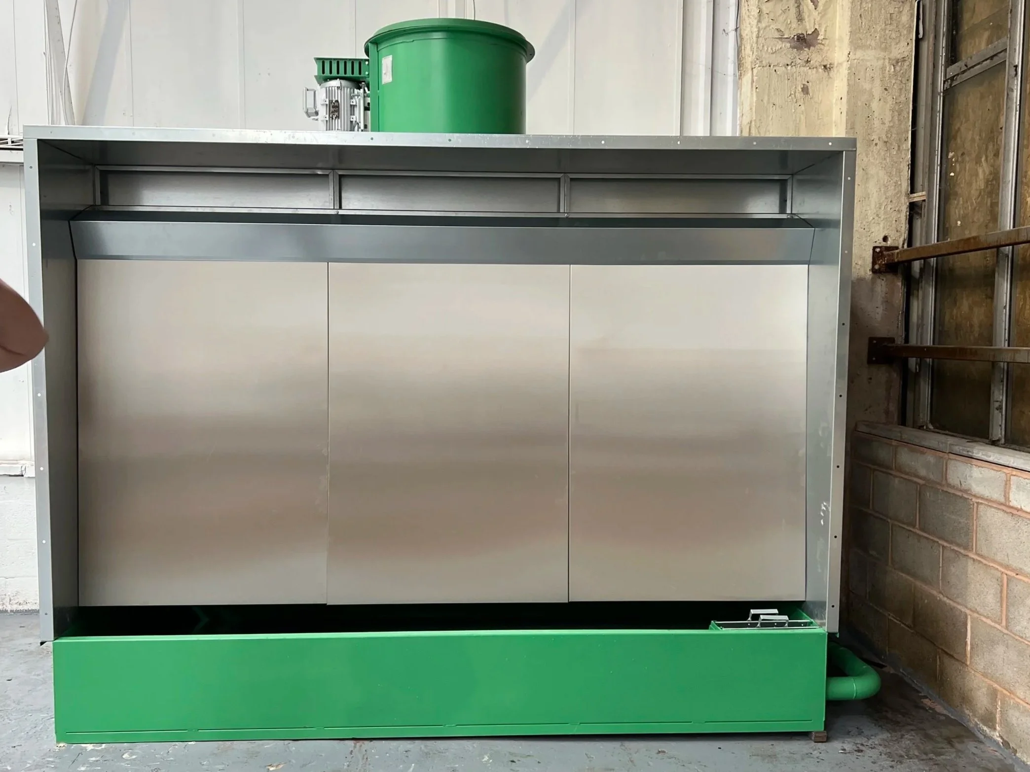 A partially assembled metal spray booth with a green base, silver top, and cabinet doors, placed against a brick and concrete wall.