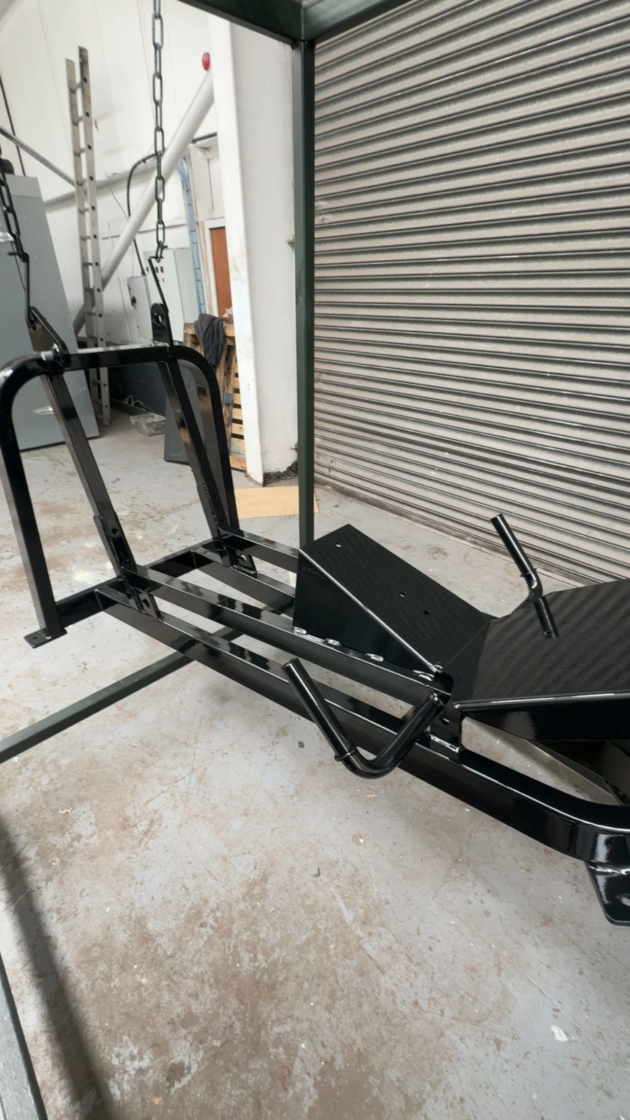 A black medical examination or therapy chair with a metal frame and adjustable headrest, situated in a workshop or storage area with concrete floor and a metal shutter door in the background.