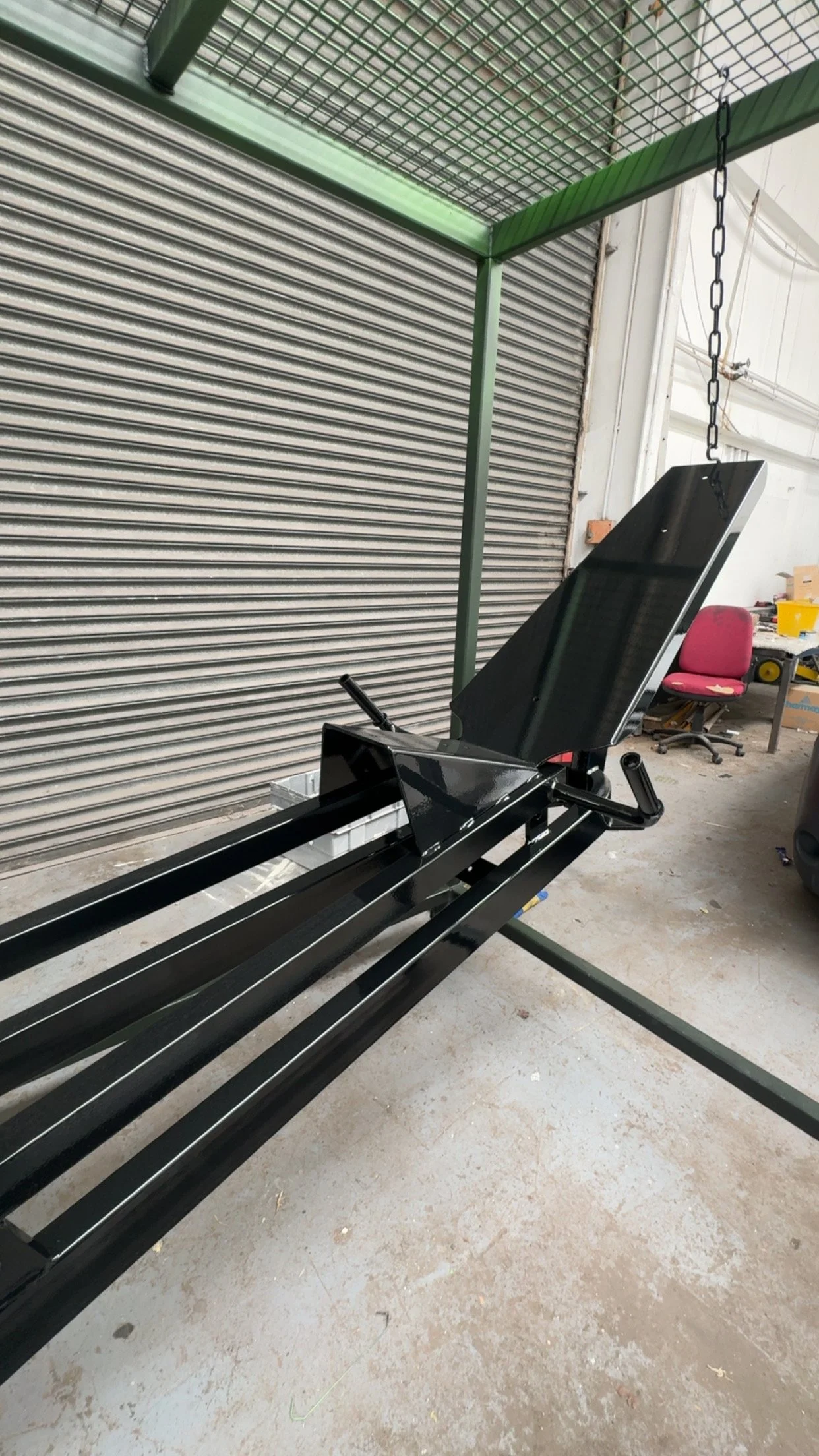 A black treadmill with a tilted screen and handrails, situated in a garage or warehouse space with concrete floor, metal shelving, a chair, and closed metal roll-up door.