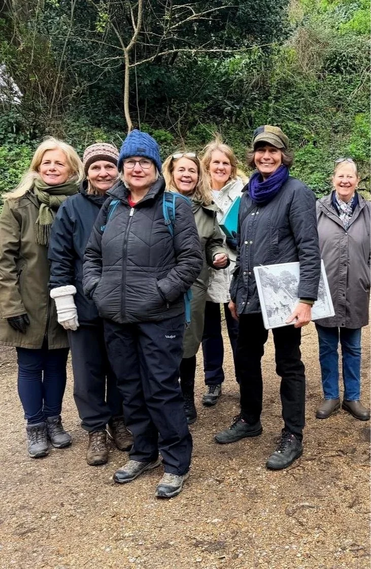 Six women dressed warmly, standing outdoors on a dirt path with greenery in the background, smiling for the camera during a group hike or outdoor gathering in cool weather.
