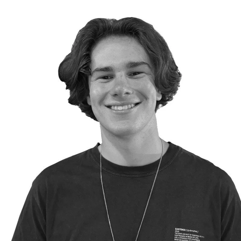 Black and white portrait of a young man with shoulder-length curly hair, smiling, wearing a black shirt and a necklace.