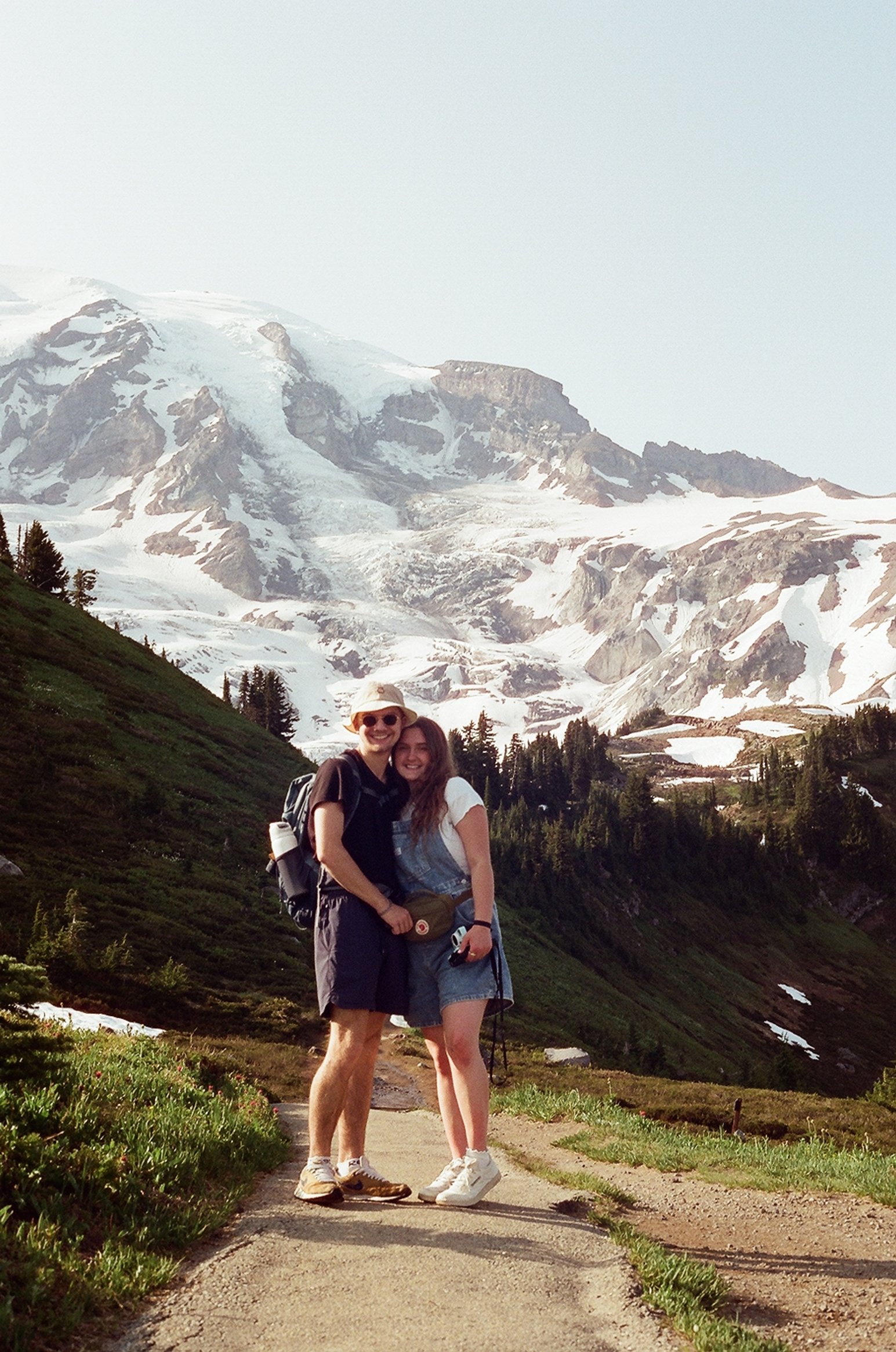 A young man and woman stand on a dirt trail in front of snow-capped mountains, smiling and hugging during a hike or outdoor adventure.