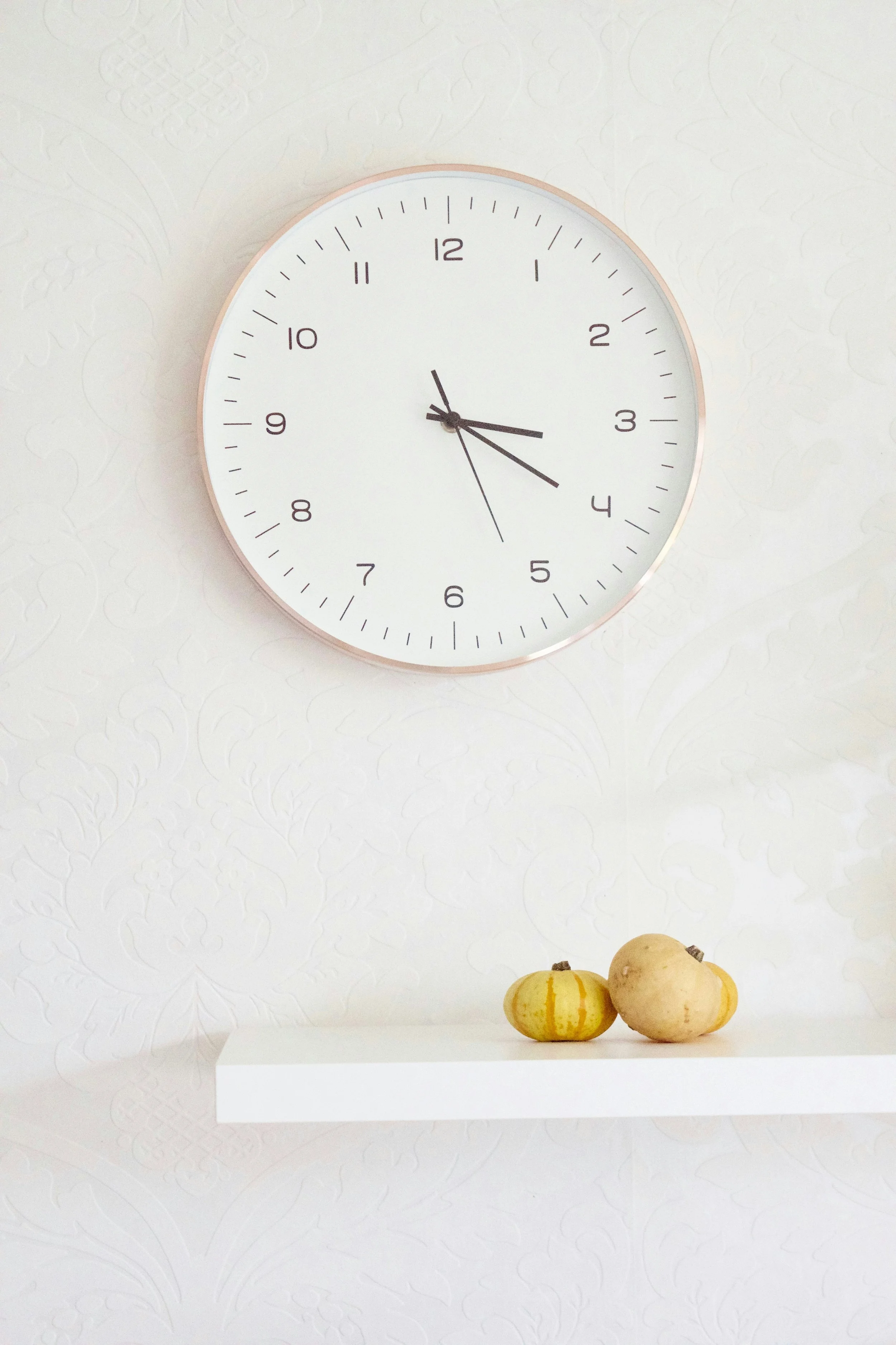 A round white wall clock showing 2:18 with a white textured background and a white shelf with three decorative gourds below it.