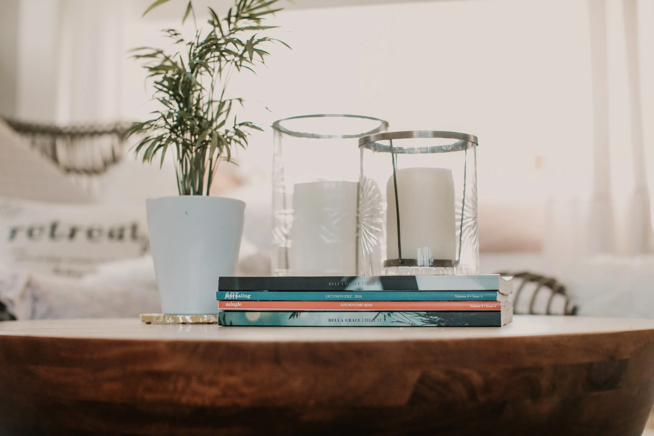 Decorative table arrangement with a potted plant, two glass candle holders with candles, and a stack of magazines on a wooden table.