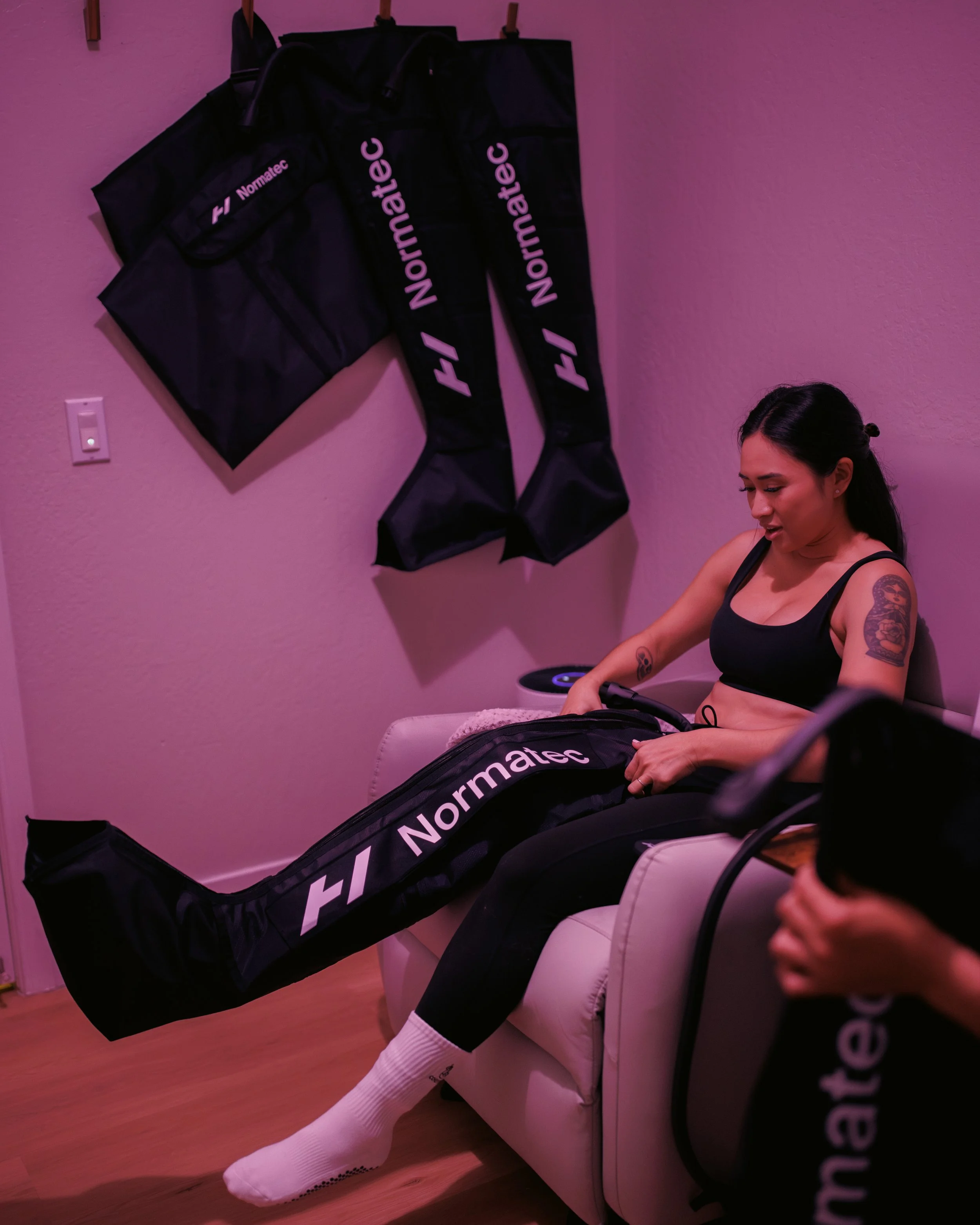 A woman sitting on a white couch with her legs in compression pants labeled 'Normatec.' She is in a room with nice lighting, with a wall-mounted set of compression sleeves and pants labeled 'Normatec' hanging on the wall behind her.