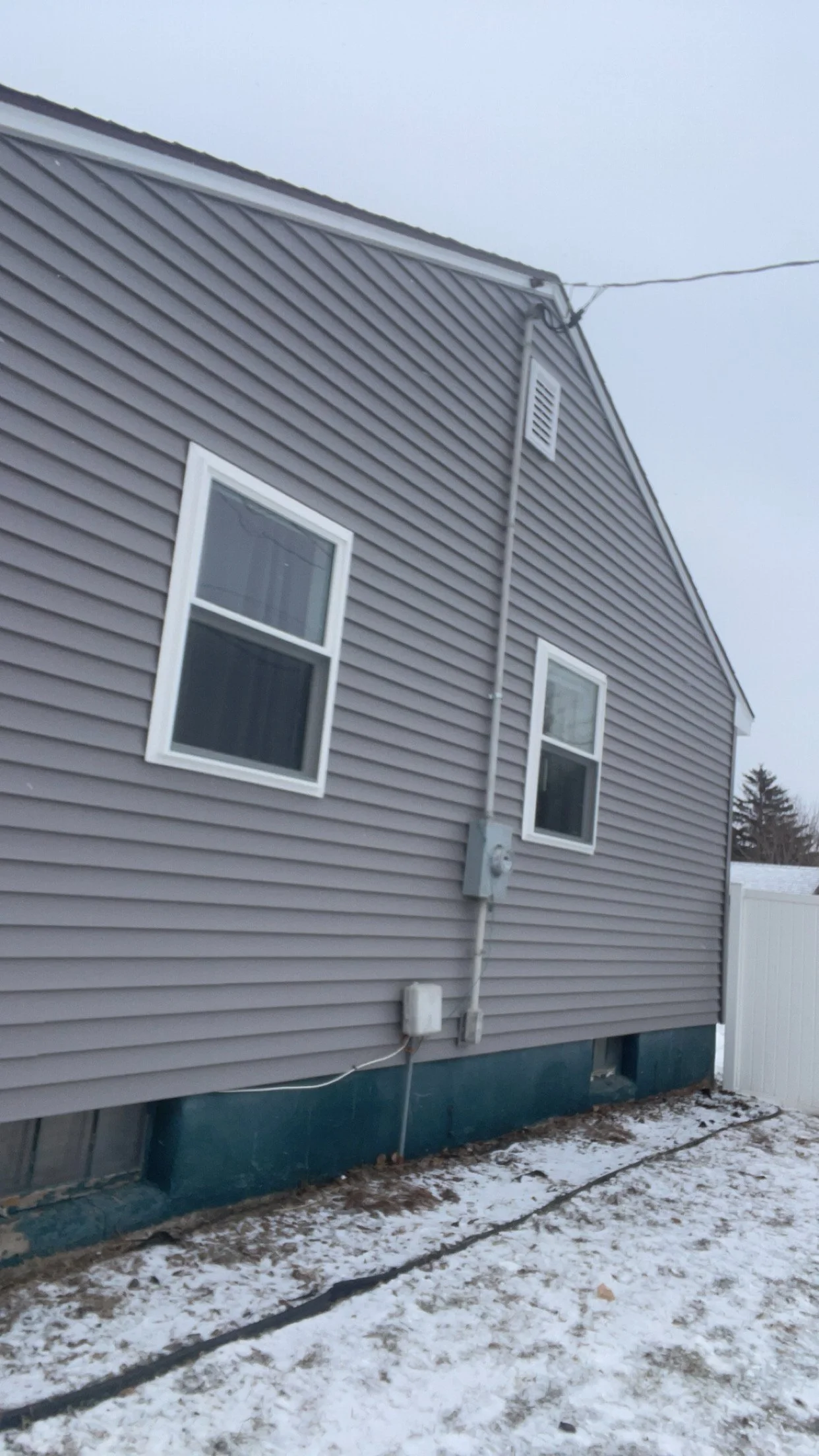 Side of a gray house with two white-framed windows, electrical meter, and utility box, with snow on the ground, overcast sky, and white fence in the background.